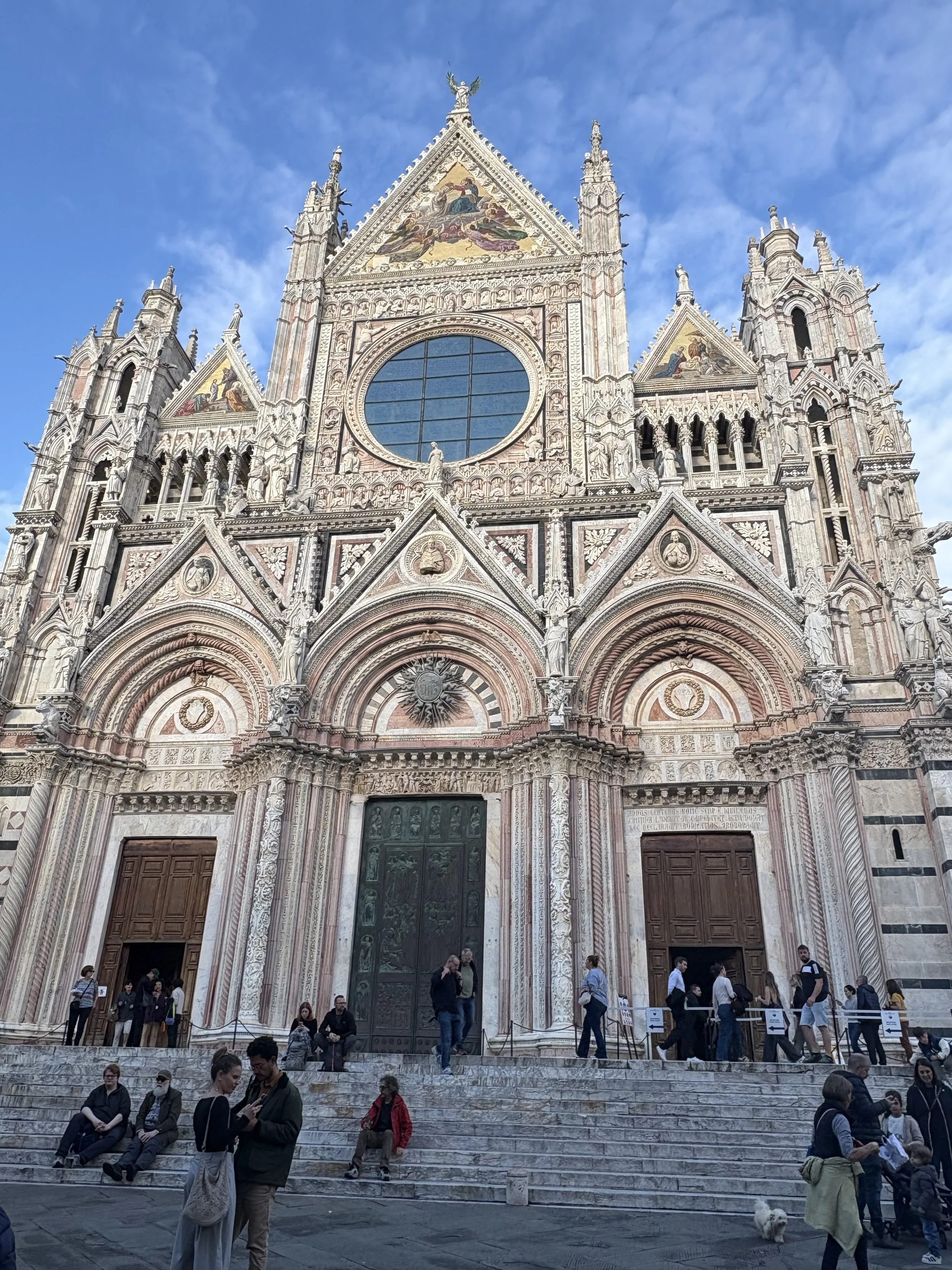Front view of the ornate Siena Cathedral in Italy with people sitting on steps and walking in front.