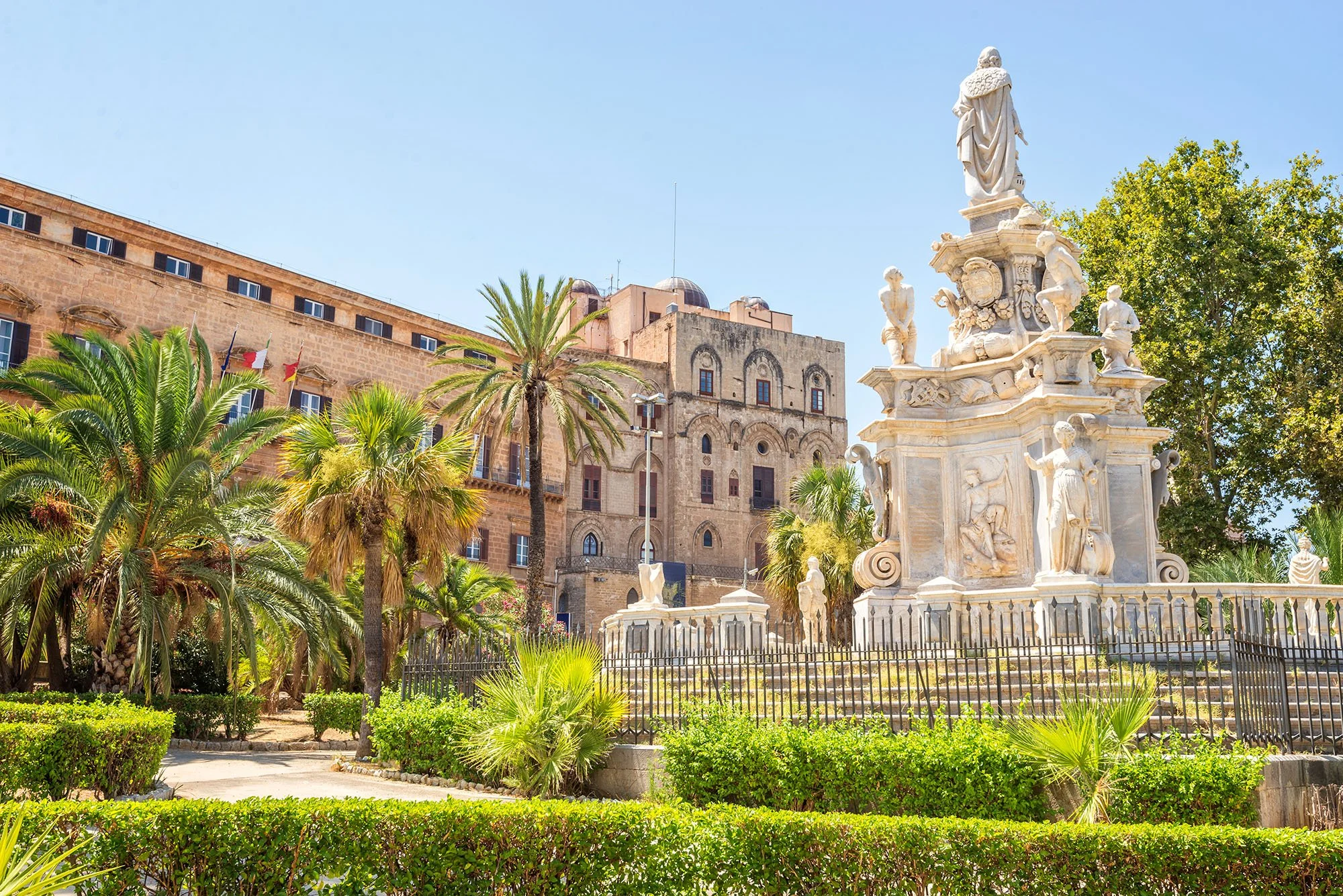A historic square with a white marble fountain and statue surrounded by green bushes and palm trees under a clear blue sky.