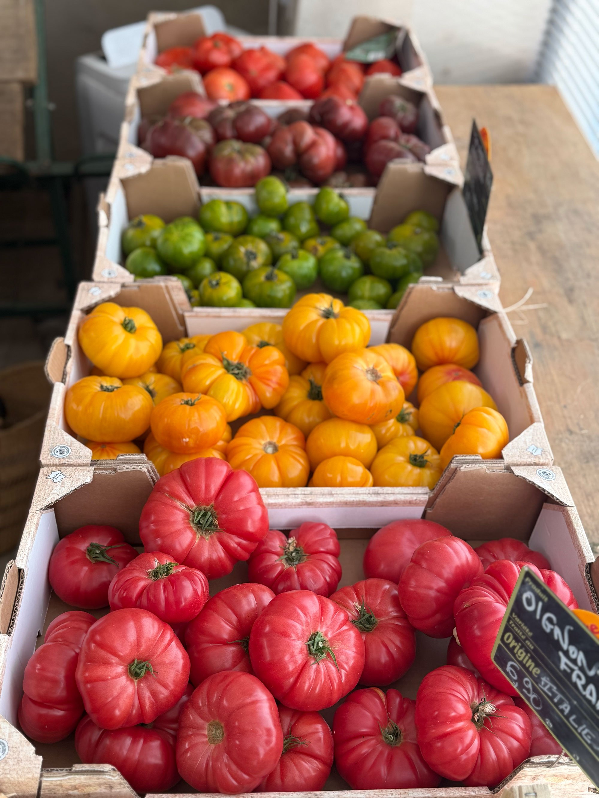Boxes of heirloom tomatoes in red, green, and yellow-orange colors displayed on a wooden table at a market.