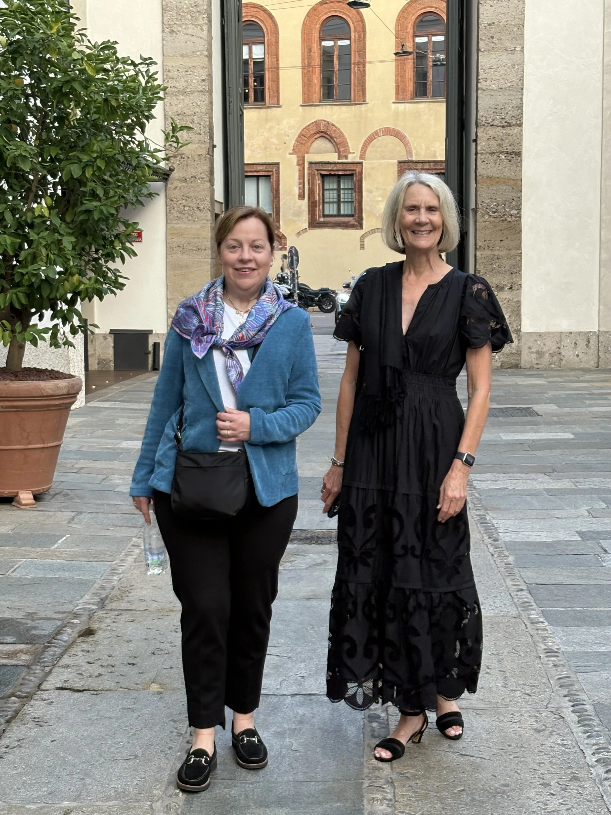 Two women standing outdoors on a cobblestone street with historic buildings in the background, smiling at the camera.
