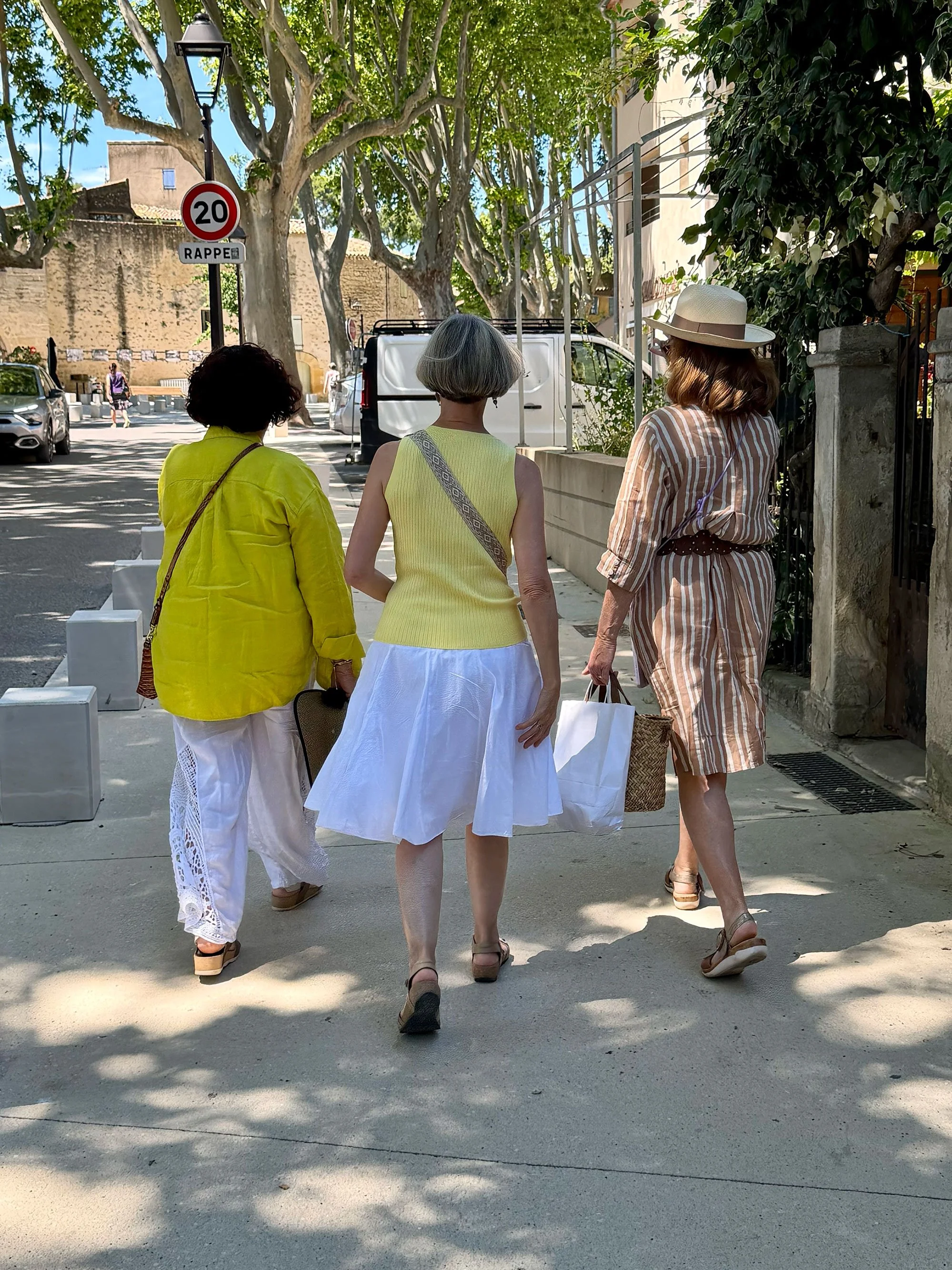Three women walking together on a street in a sunny day, wearing colorful summer dresses and hats, with trees and parked cars in the background.