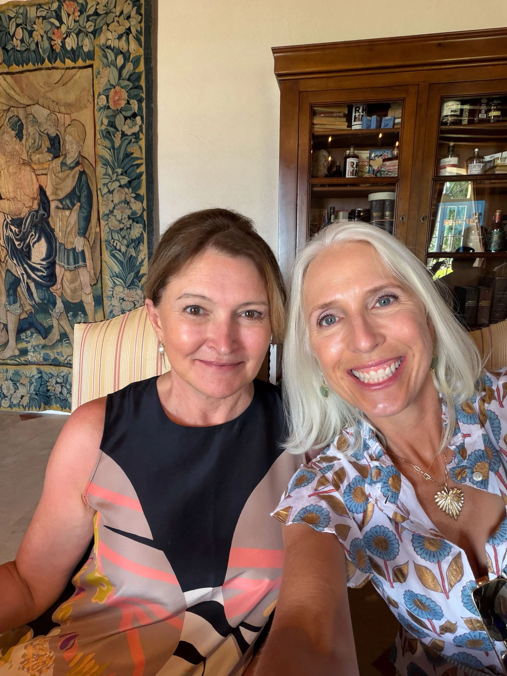 Two smiling women taking a selfie indoors with a tapestry and wooden cabinet in the background.
