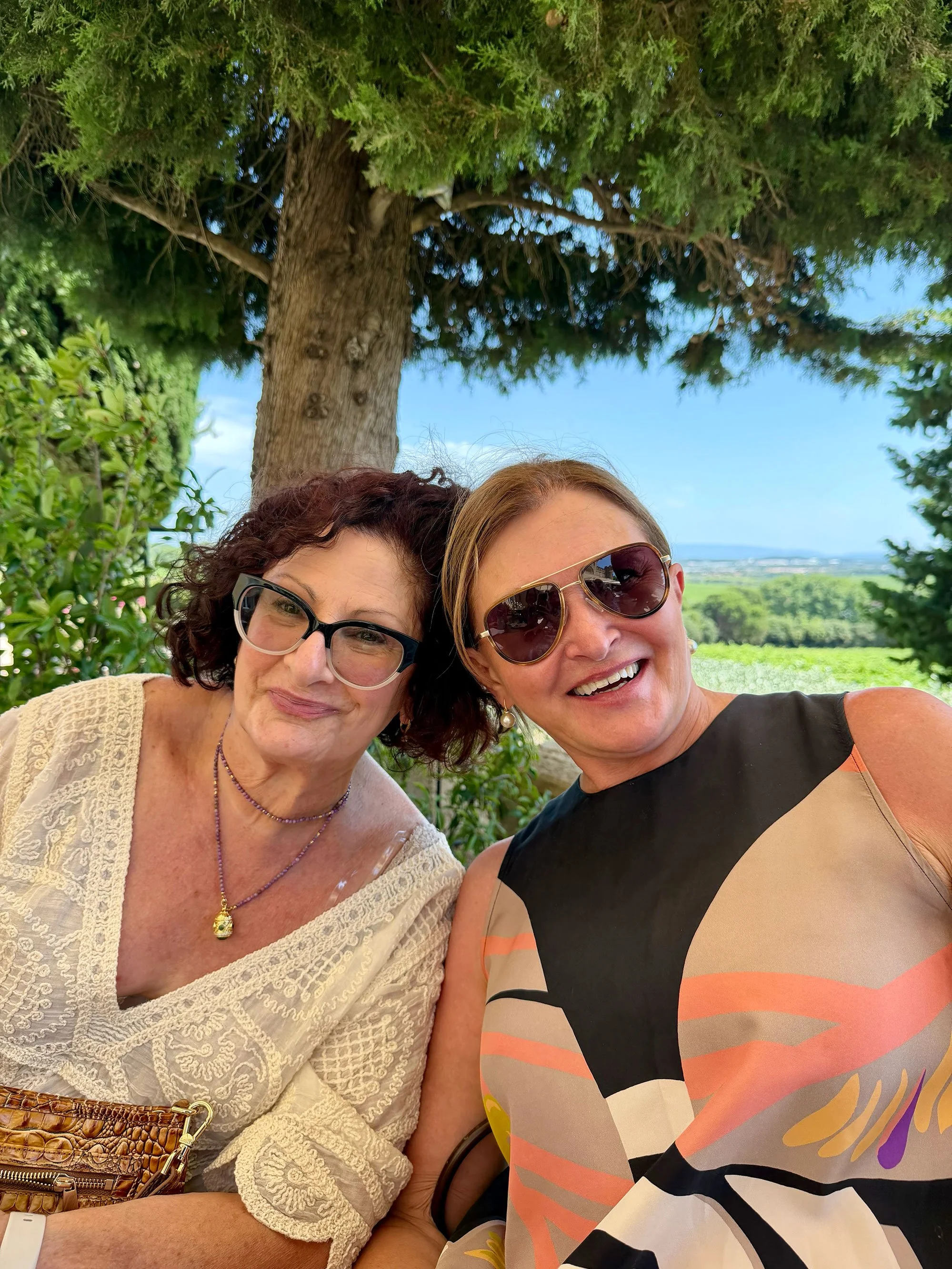 Two women smiling and taking a selfie outdoors with a large tree and open landscape in the background. They are wearing sunglasses and casual clothing.