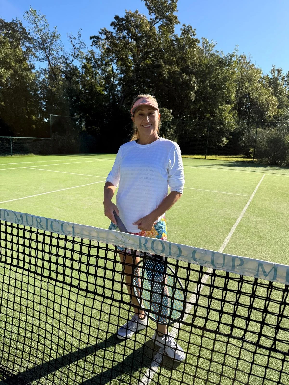 A woman standing on a tennis court holding a tennis racket, smiling, wearing a pink visor, white long sleeve shirt, colorful shorts, and white tennis shoes, with trees and a clear blue sky in the background.