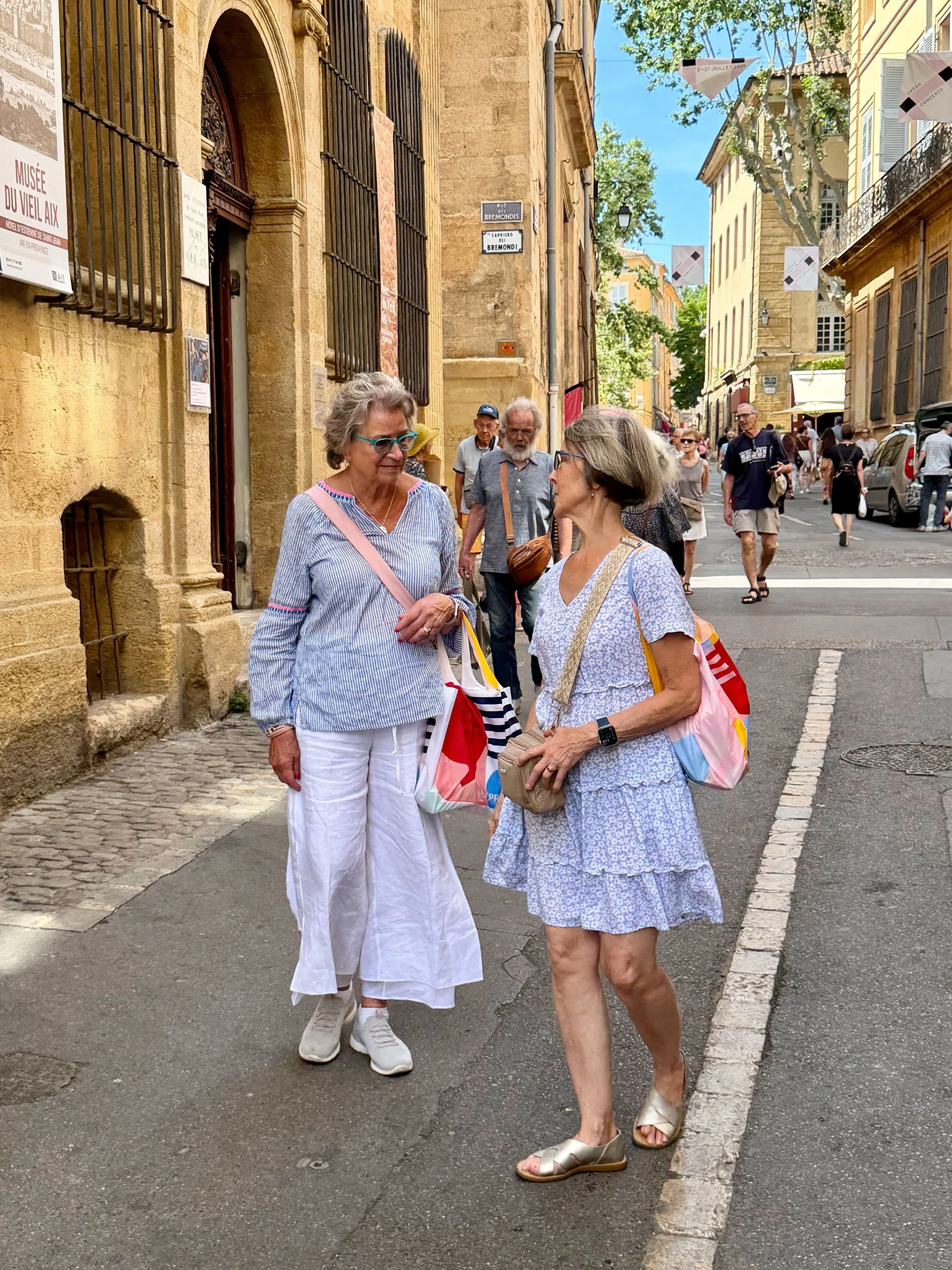 Two women talking on a city street with other pedestrians and buildings in the background.