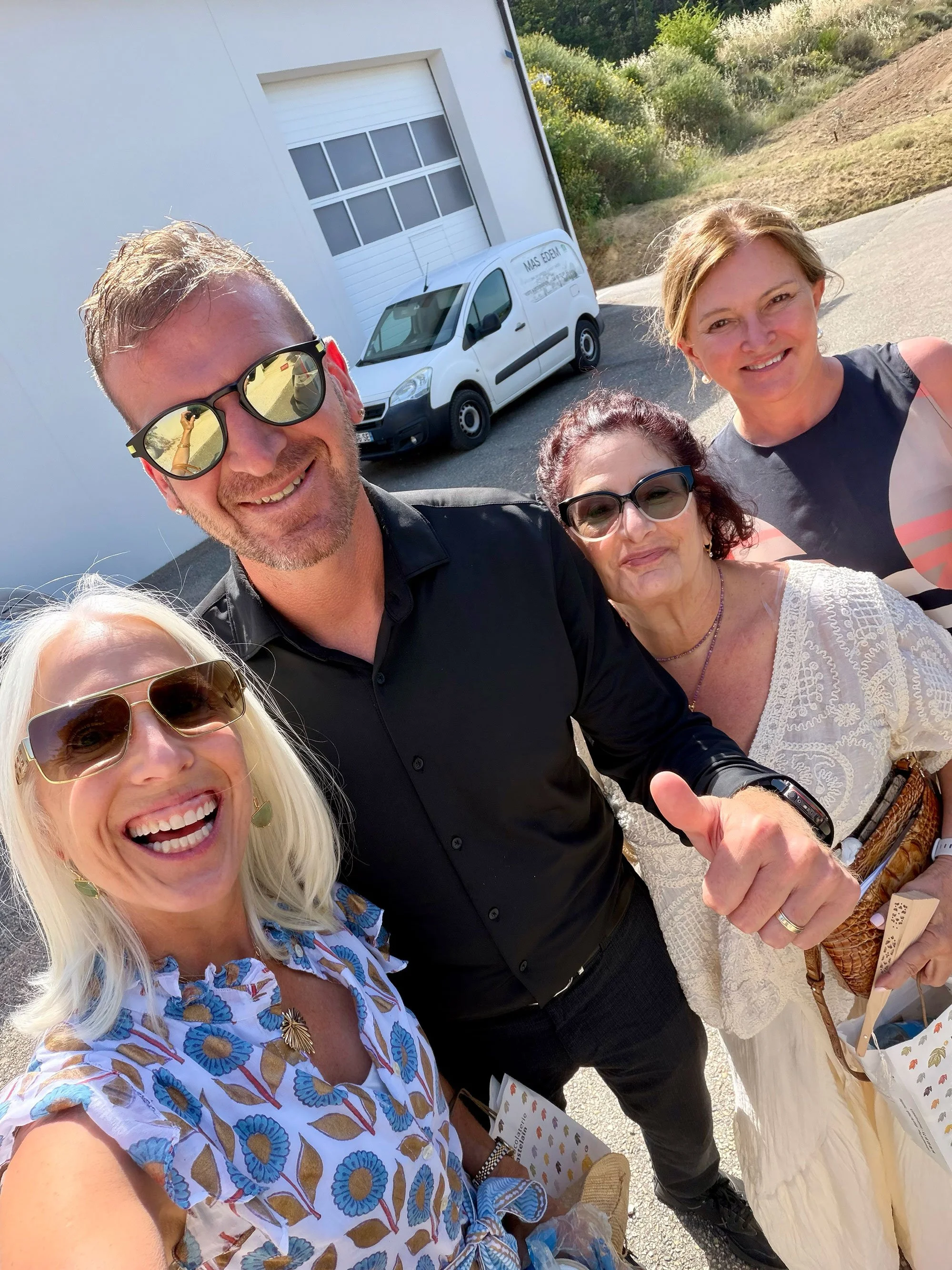 Group selfie of four smiling people, three women and one man, outdoors on a sunny day. One woman wears sunglasses and a floral dress; another woman wears glasses and a cream lace top; the third woman has light hair and wears a sleeveless top. The man