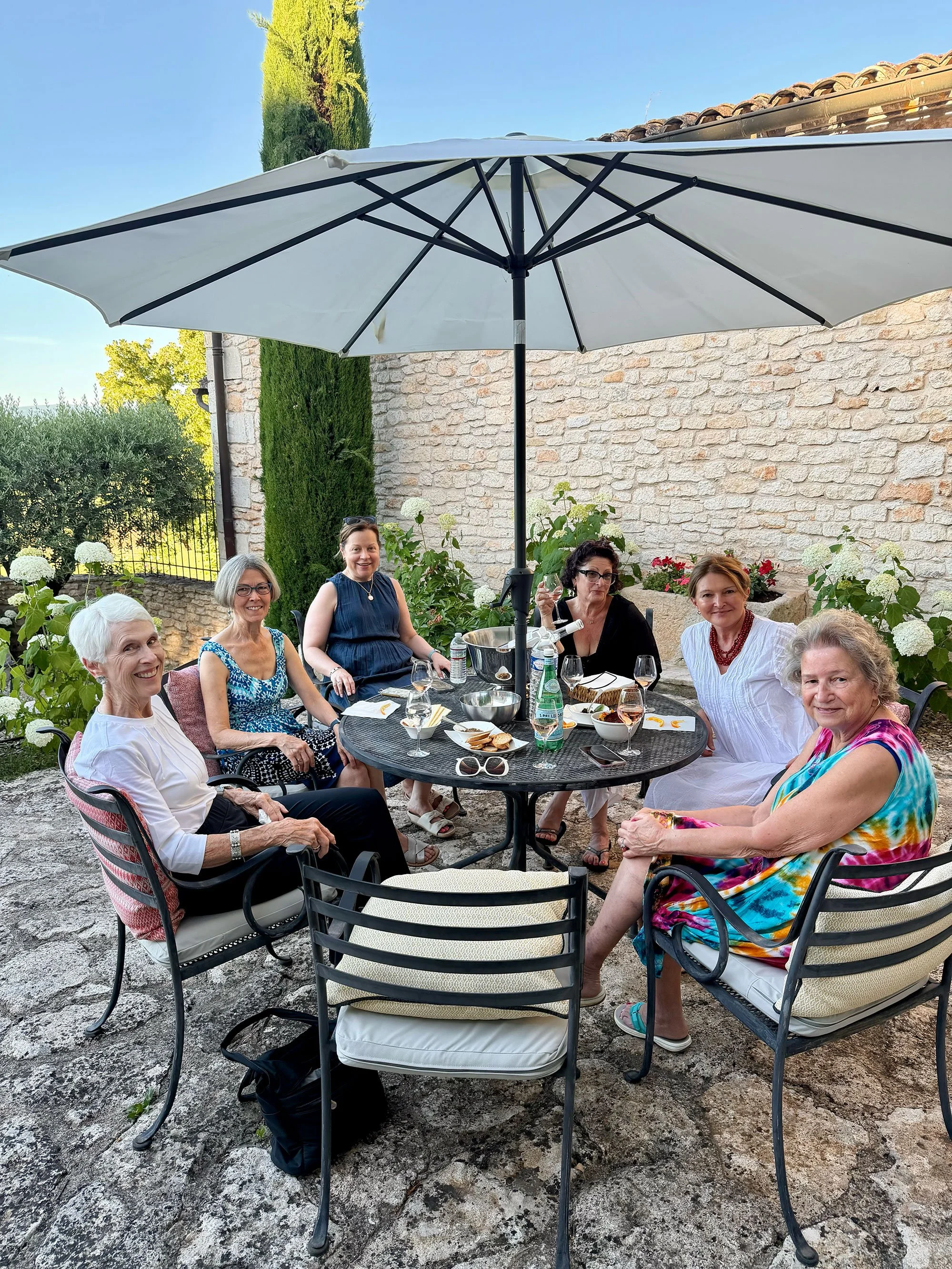 A group of seven women sitting around a black outdoor table under a large white patio umbrella, enjoying drinks and food in a garden setting with green plants, flowers, and a brick wall behind them.