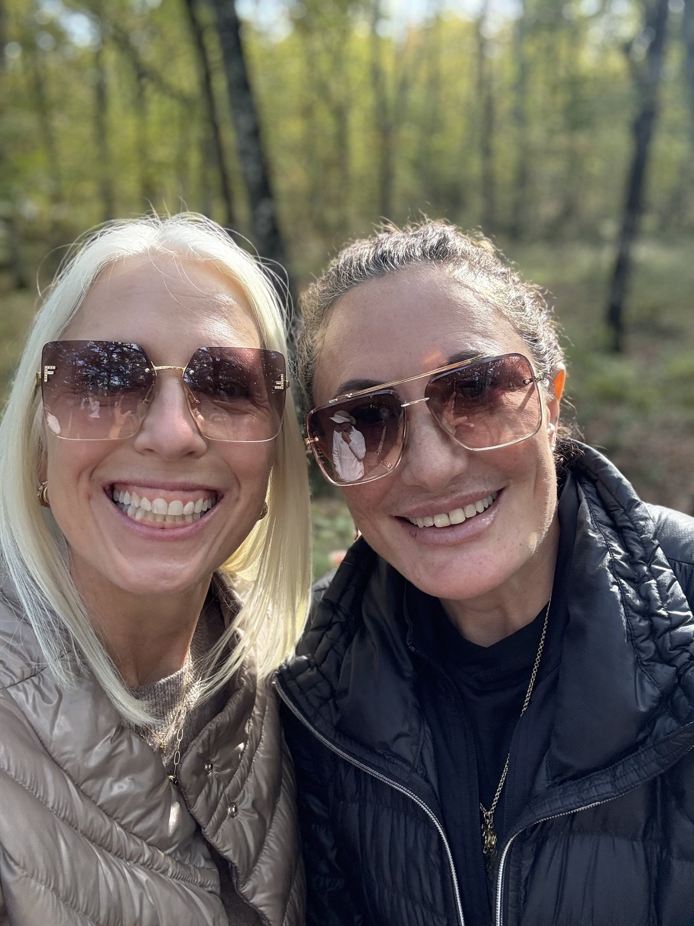 Two smiling women wearing sunglasses taking a selfie outdoors in a wooded area during daylight.