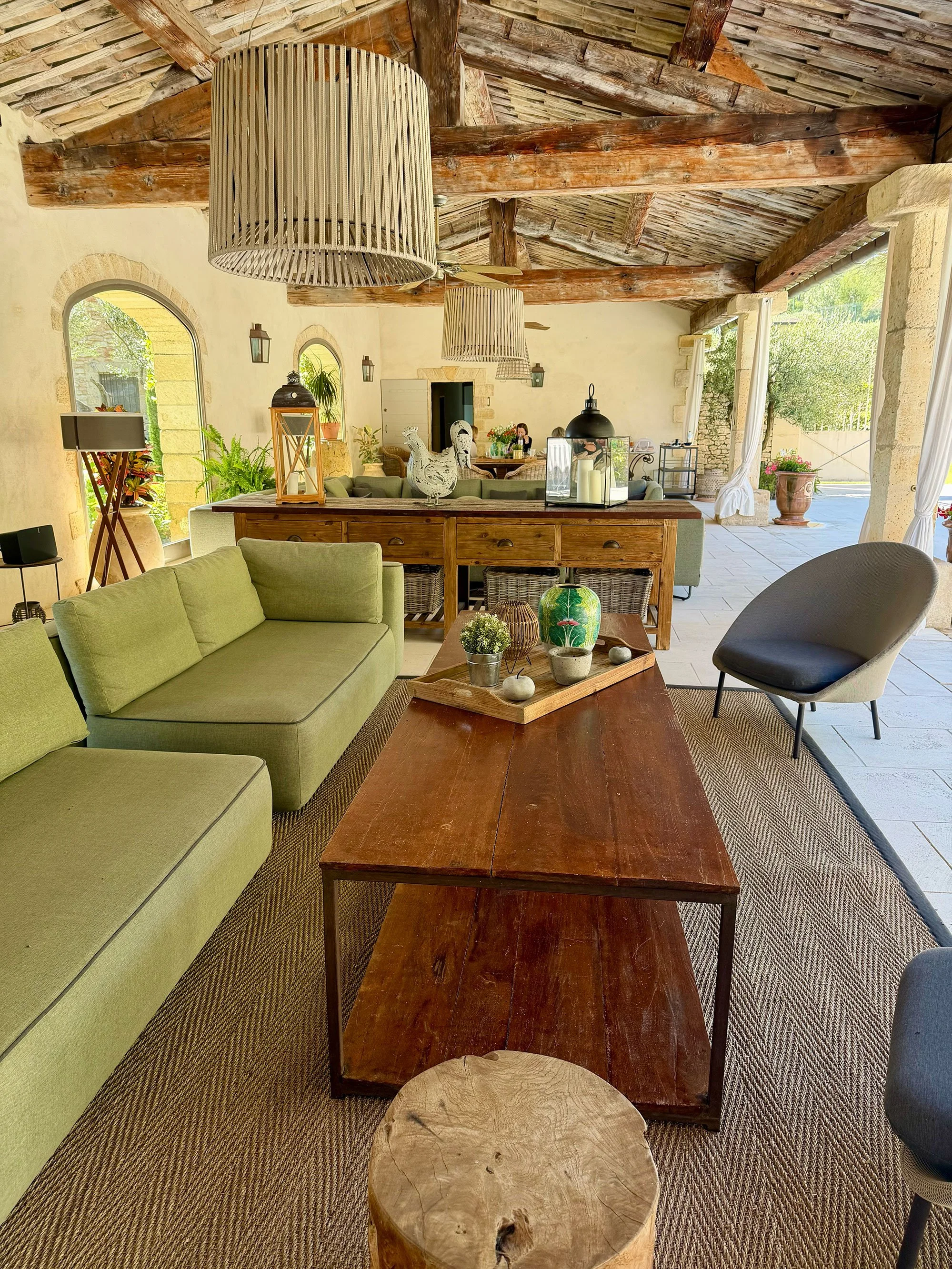 Living room with wooden beams on the ceiling, green sofa, various decorative plants, tables, and large outdoor windows.