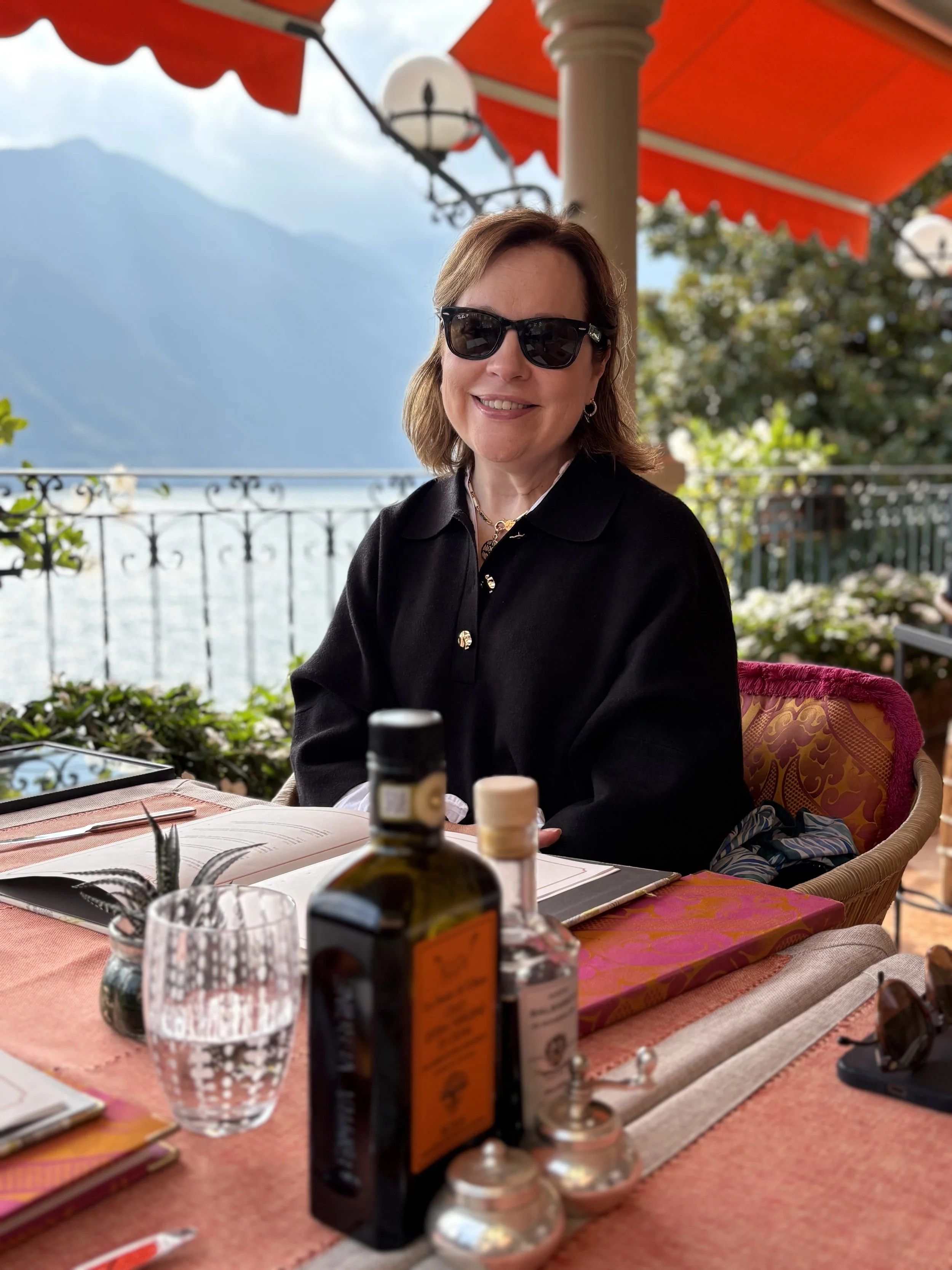 A woman sitting at an outdoor restaurant table near a lake, wearing sunglasses and a black top. The table has a water glass, bottles, and menus. The background features mountains, a railing, and red awnings.