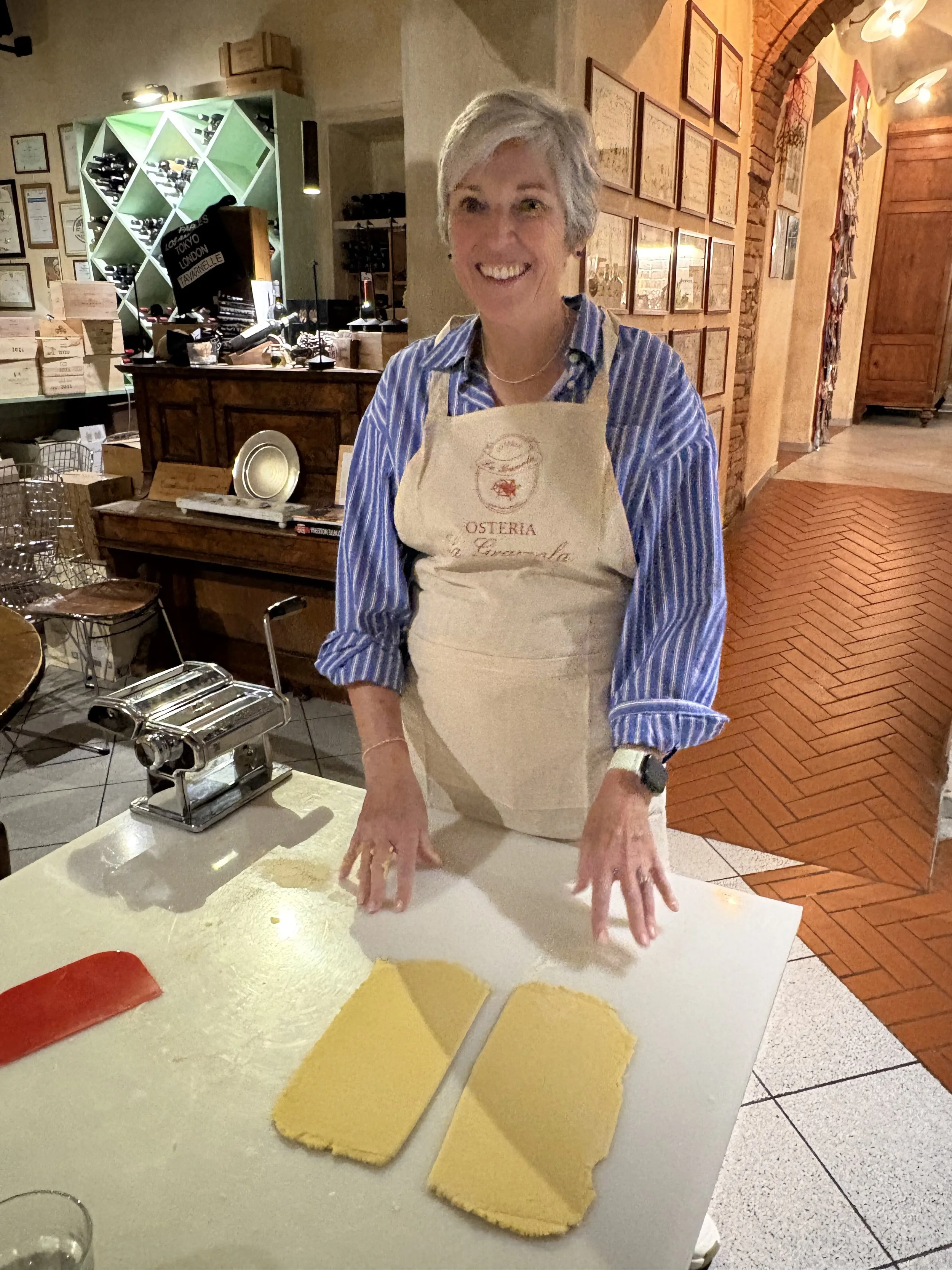 A woman with short gray hair smiling and wearing a striped blue shirt and a beige apron, standing at a table with two slices of yellow cake or pastry, and a red knife, inside a cozy restaurant or bakery.