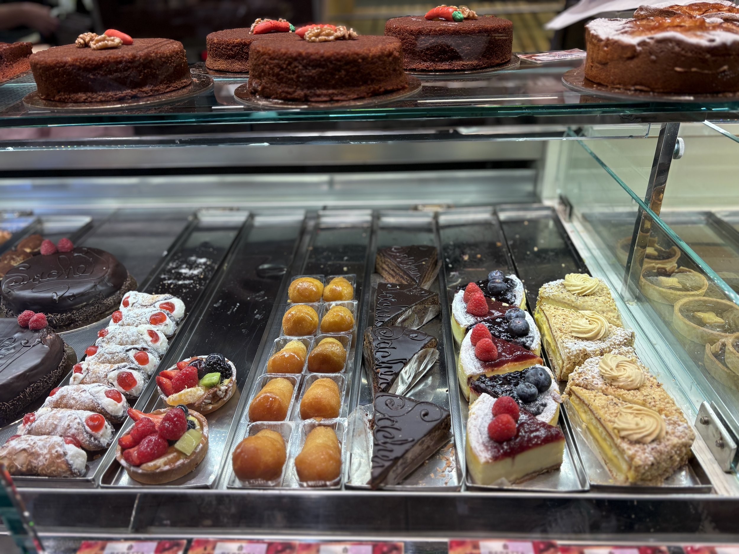 Display case with various cakes and pastries, including chocolate cakes, fruit-topped cheesecakes, and small decorated pastries topped with fruits and powdered sugar.