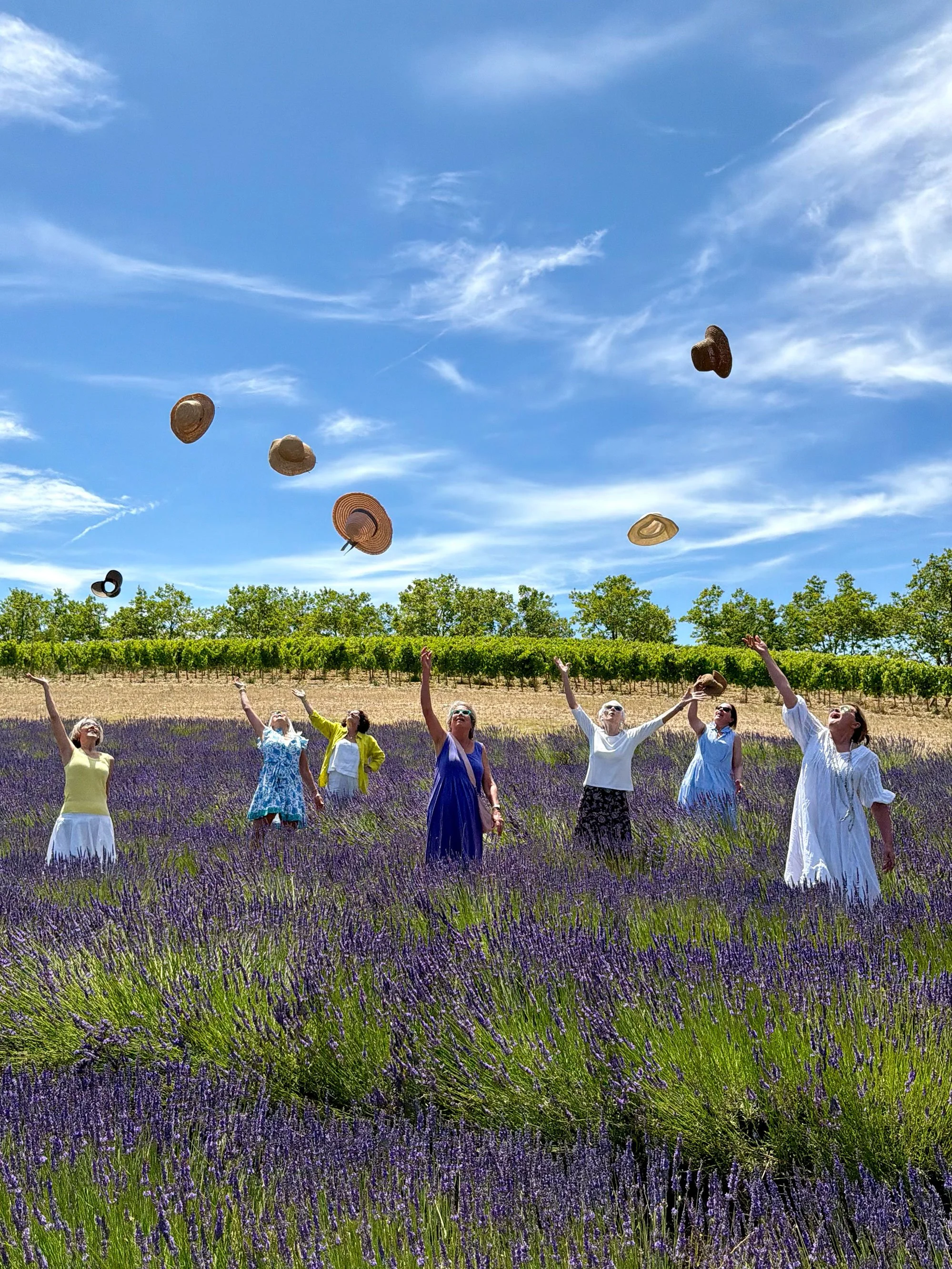 A group of women in colorful dresses stand in a lavender field, tossing hats into the air under a blue sky with wispy clouds.