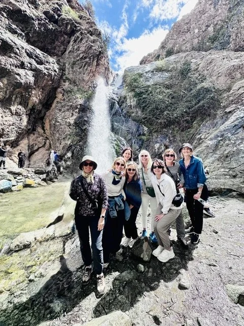Group of women and men hiking near a waterfall in a rocky, mountainous area with clear water and a partly cloudy sky.