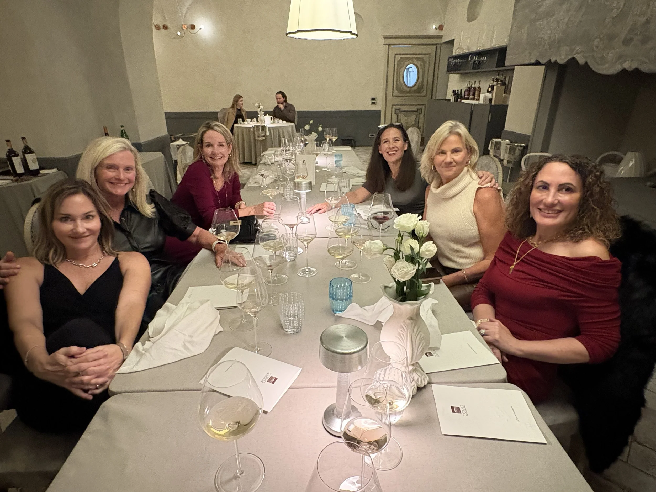 Six women sitting at a table in a restaurant, smiling, with wine glasses and a flower arrangement in the center.
