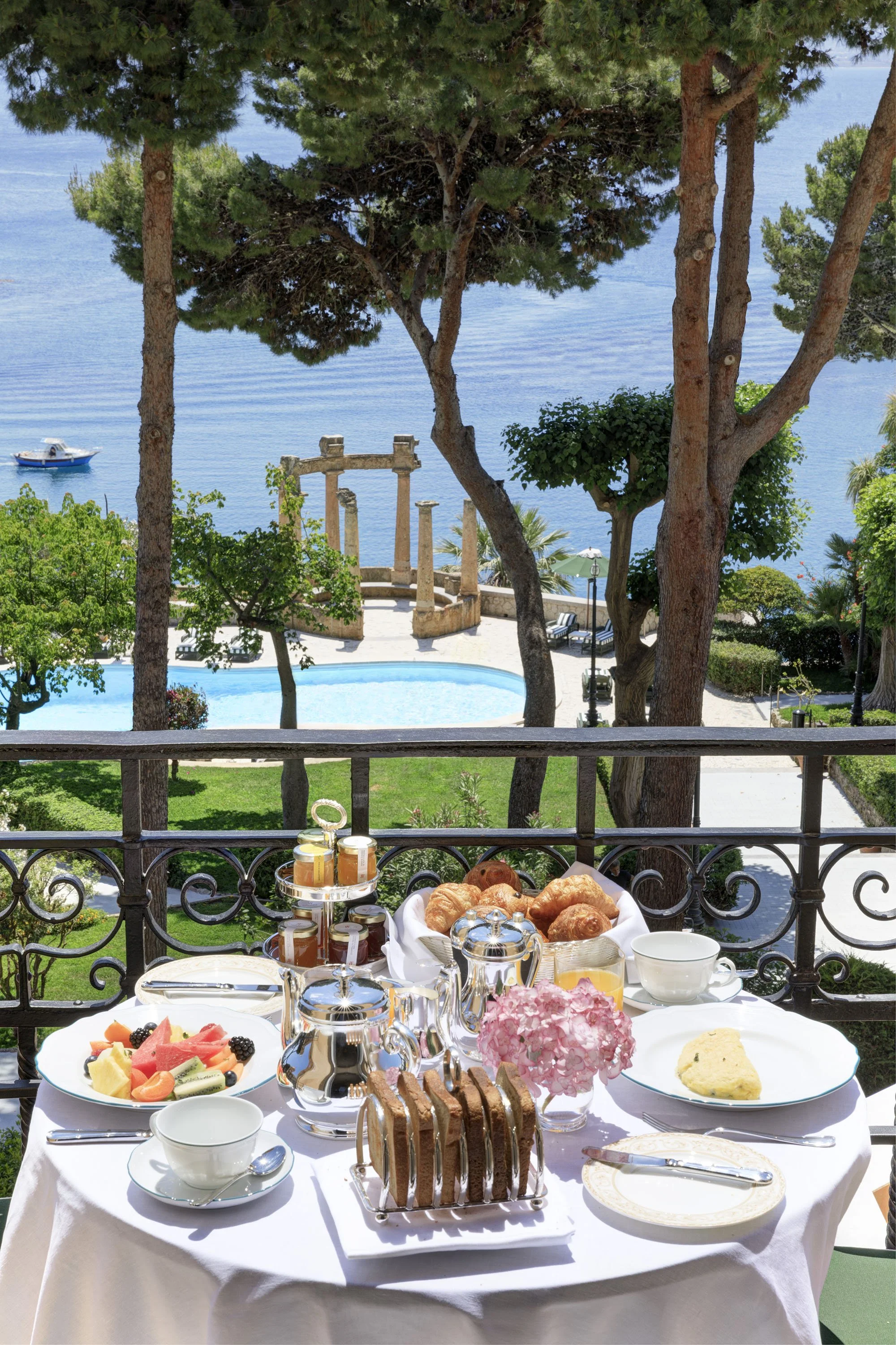 A breakfast table set outdoors with a view of the ocean, trees, and a boat. The table has various dishes including croissants, fresh fruit, pink flowers, and jars of jam, with a scenic ocean backdrop.