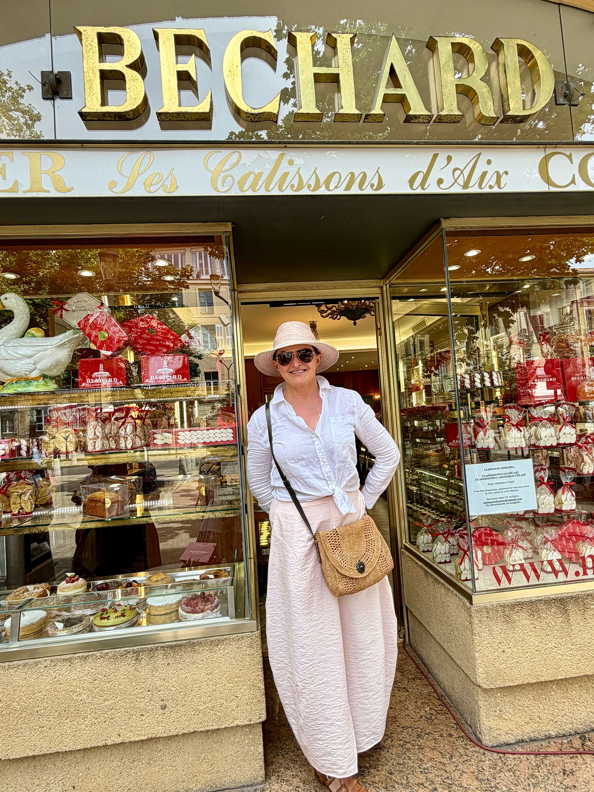 Woman wearing a white hat, sunglasses, white blouse, and pink skirt standing outside a bakery with a display case of cakes and pastries. The bakery has a sign reading 'BECHARD' and decorative elements with red ribbons and holiday-themed items.