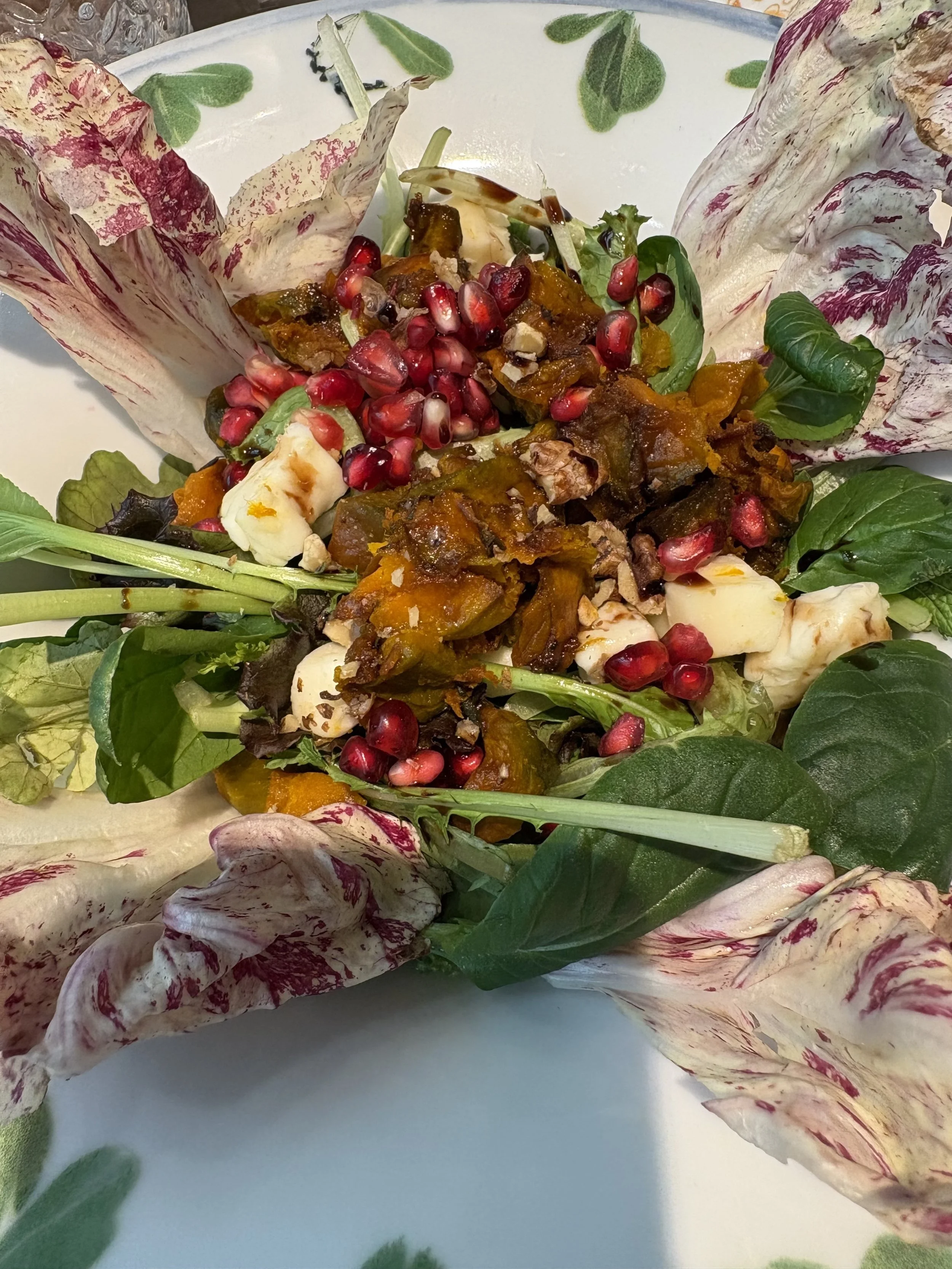 A fresh salad served on a white plate with leafy greens, pomegranate seeds, cheese, caramelized onions, and radicchio leaves arranged around the edges.