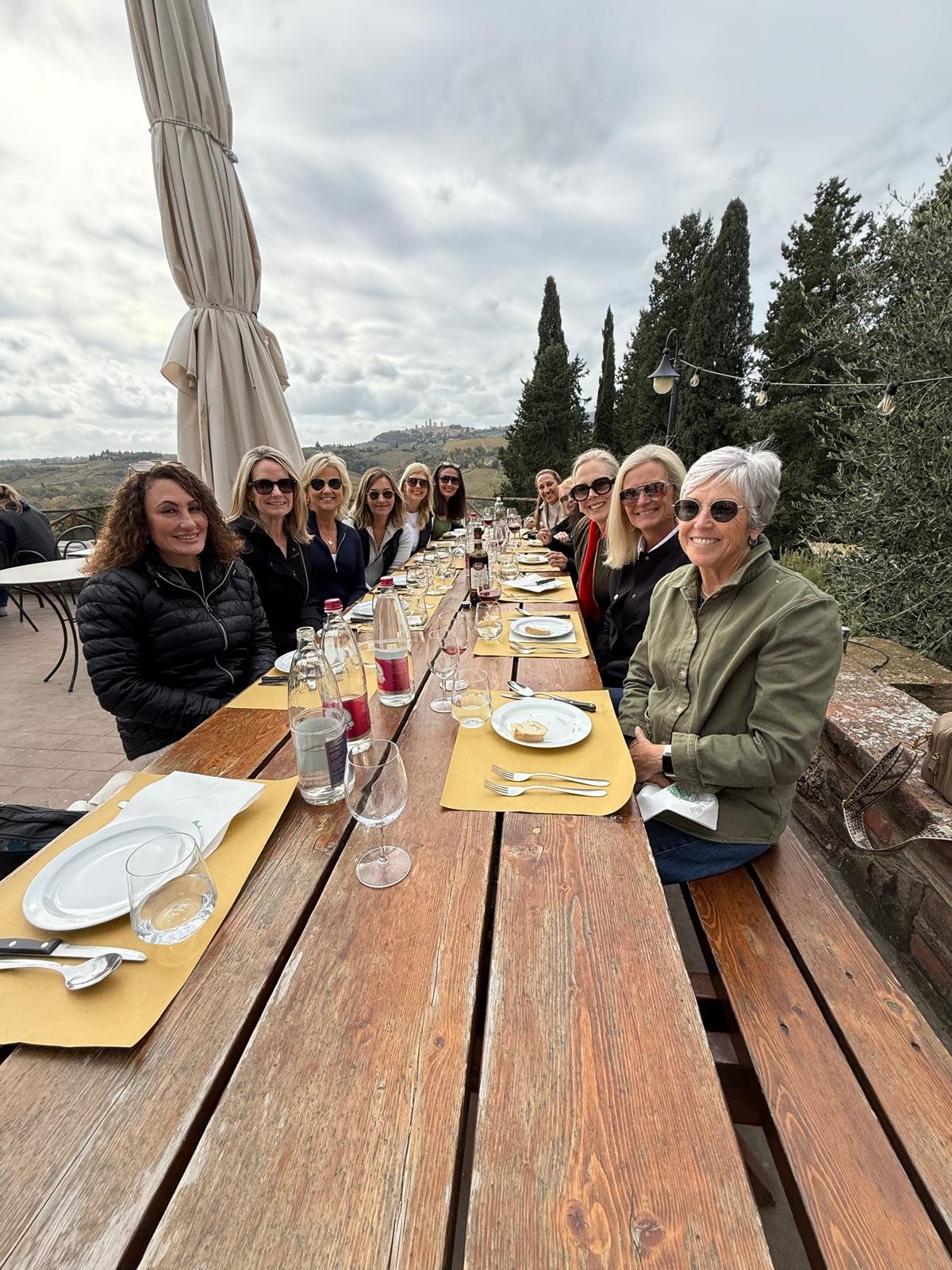 Group of women sitting at an outdoor restaurant table with plates, glasses, and bottles, smiling for the camera against a scenic backdrop of trees and hills.