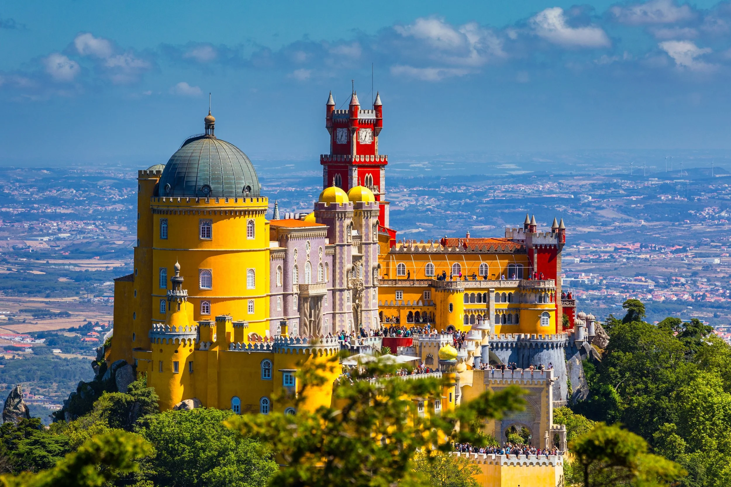 Colorful fairy tale castle with yellow, red, and purple towers on a hilltop, surrounded by green trees, under a partly cloudy sky.
