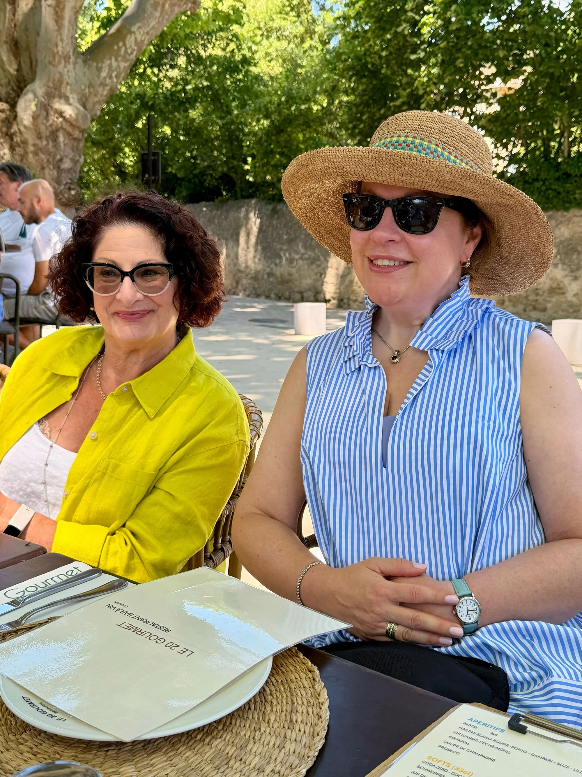 Two women seated outdoors in a shaded area, dressed in summer clothing, wearing sunglasses and hats, smiling at the camera. There is a menu and cutlery on the table in front of them.