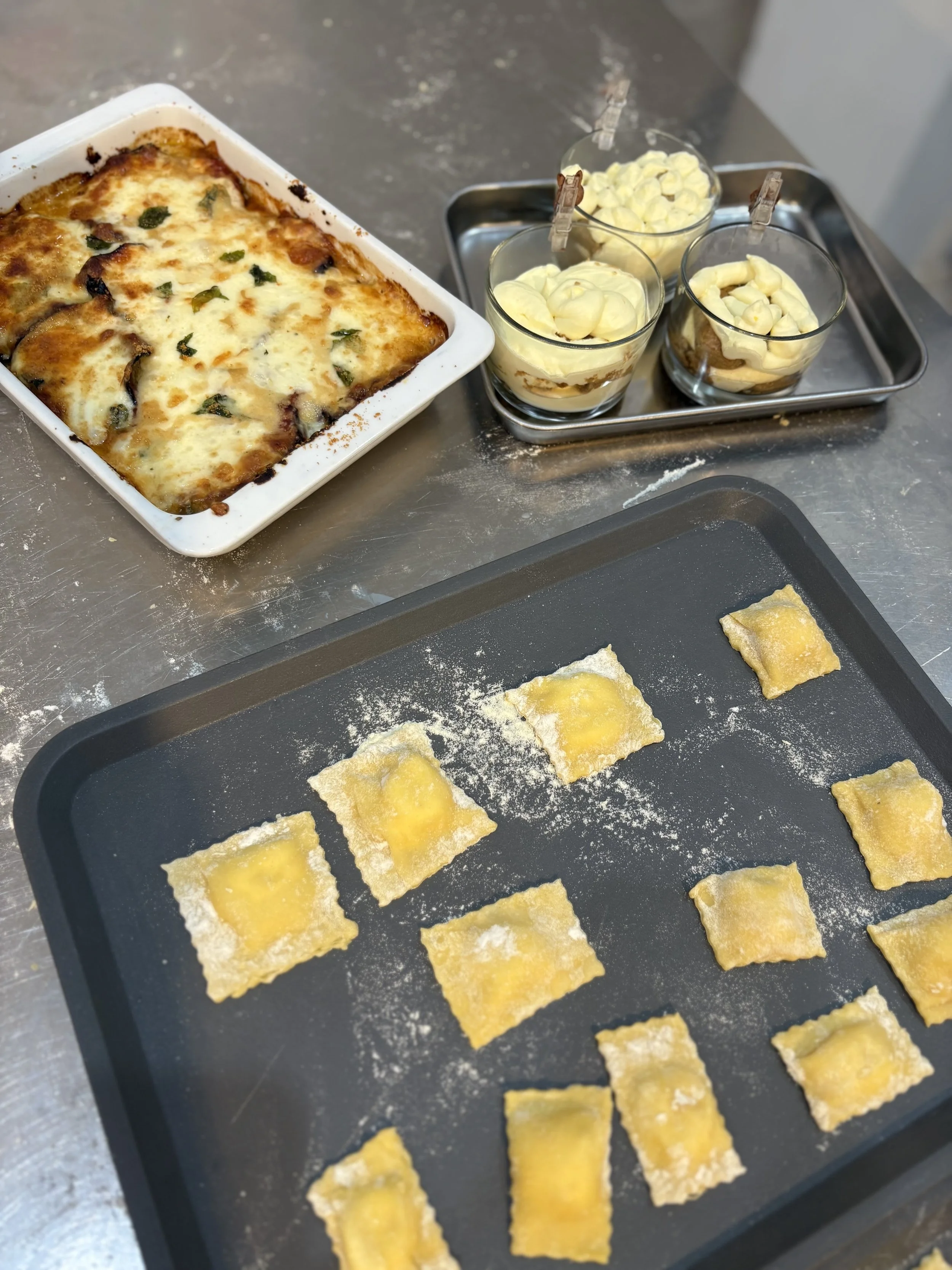 Homemade ravioli on a baking sheet in progress, with ingredients and a baked lasagna dish in the background.