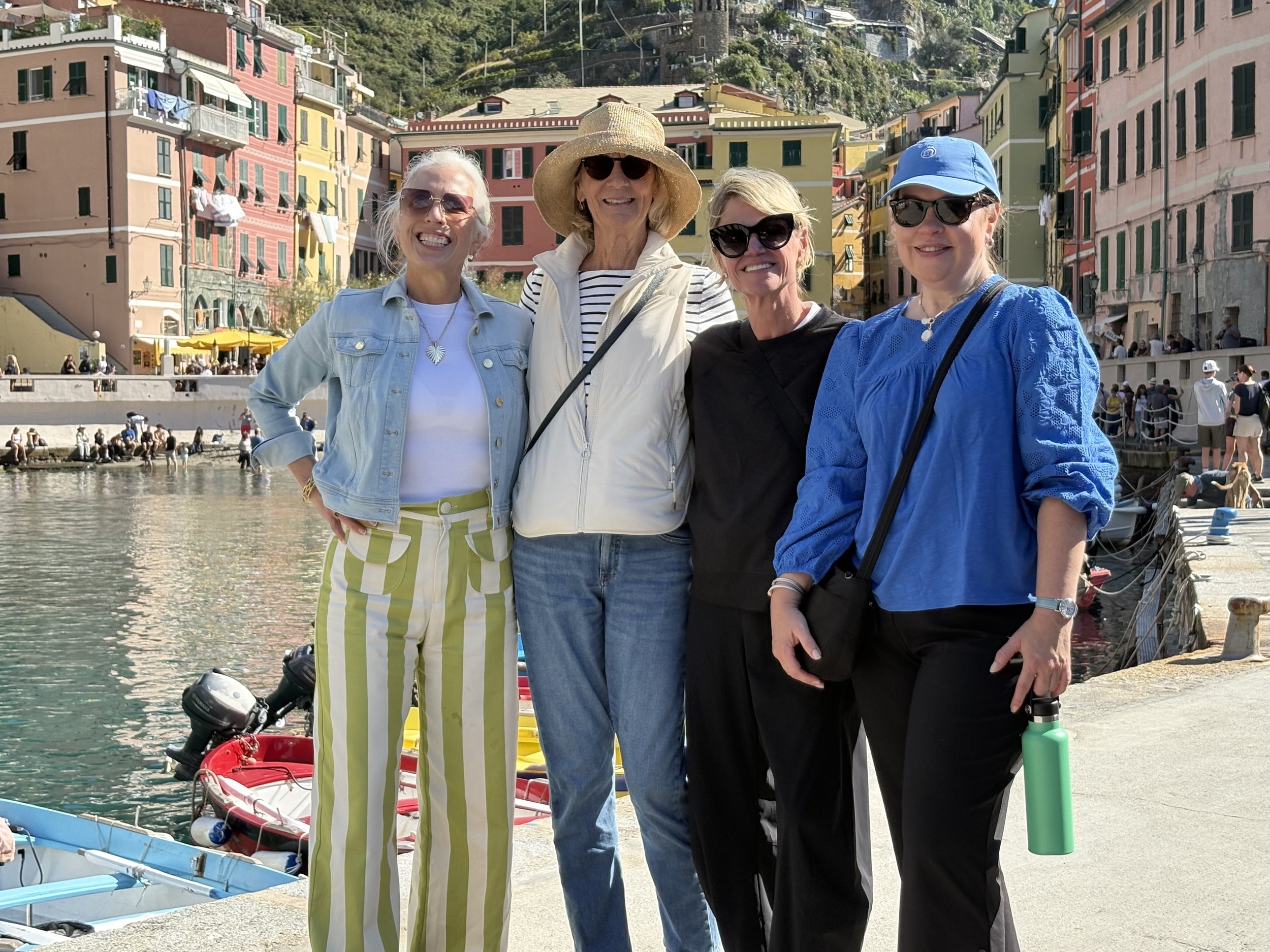 Four women standing by a waterfront with colorful buildings in the background. They are dressed casually, smiling, and wearing sunglasses, with some holding water bottles.