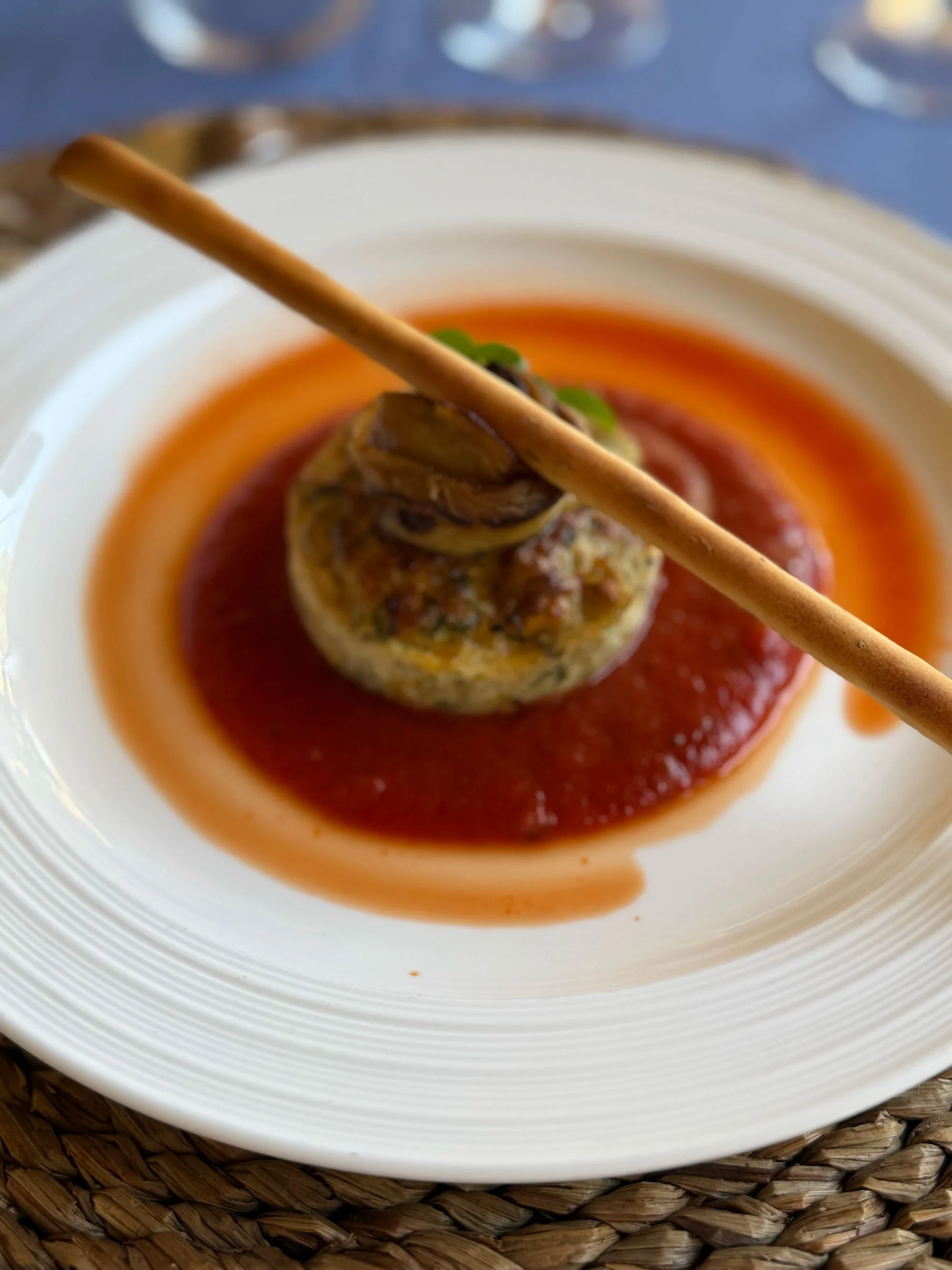 Close-up of a stuffed mushroom cap garnished with a mint leaf, resting in red sauce on a white plate. A breadstick pierces the mushroom vertically, and the plate is on a woven placemat.