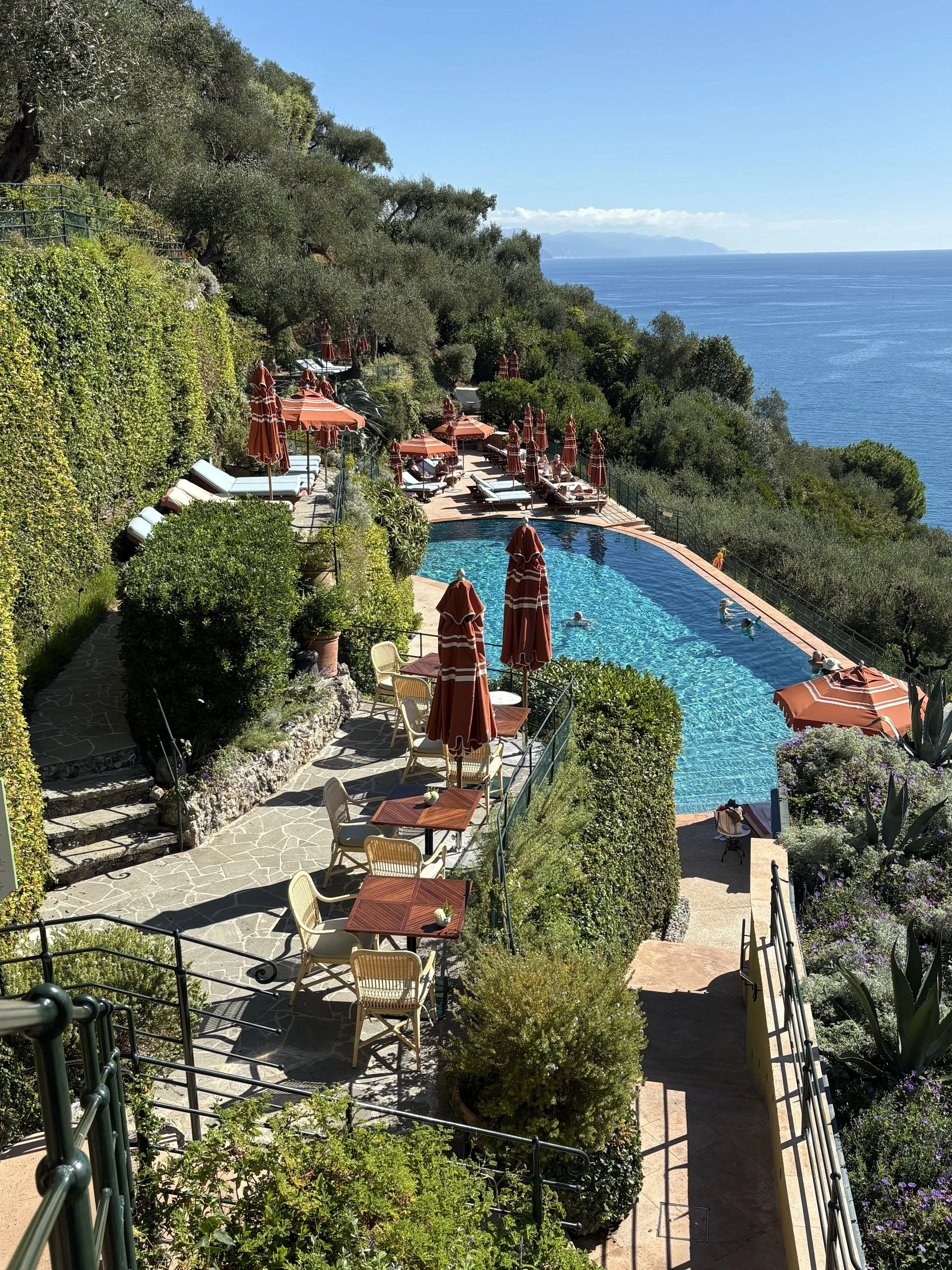 A scenic view of a swimming pool surrounded by outdoor seating with tables, chairs, and umbrellas, overlooking lush green trees and the ocean on a clear sunny day.