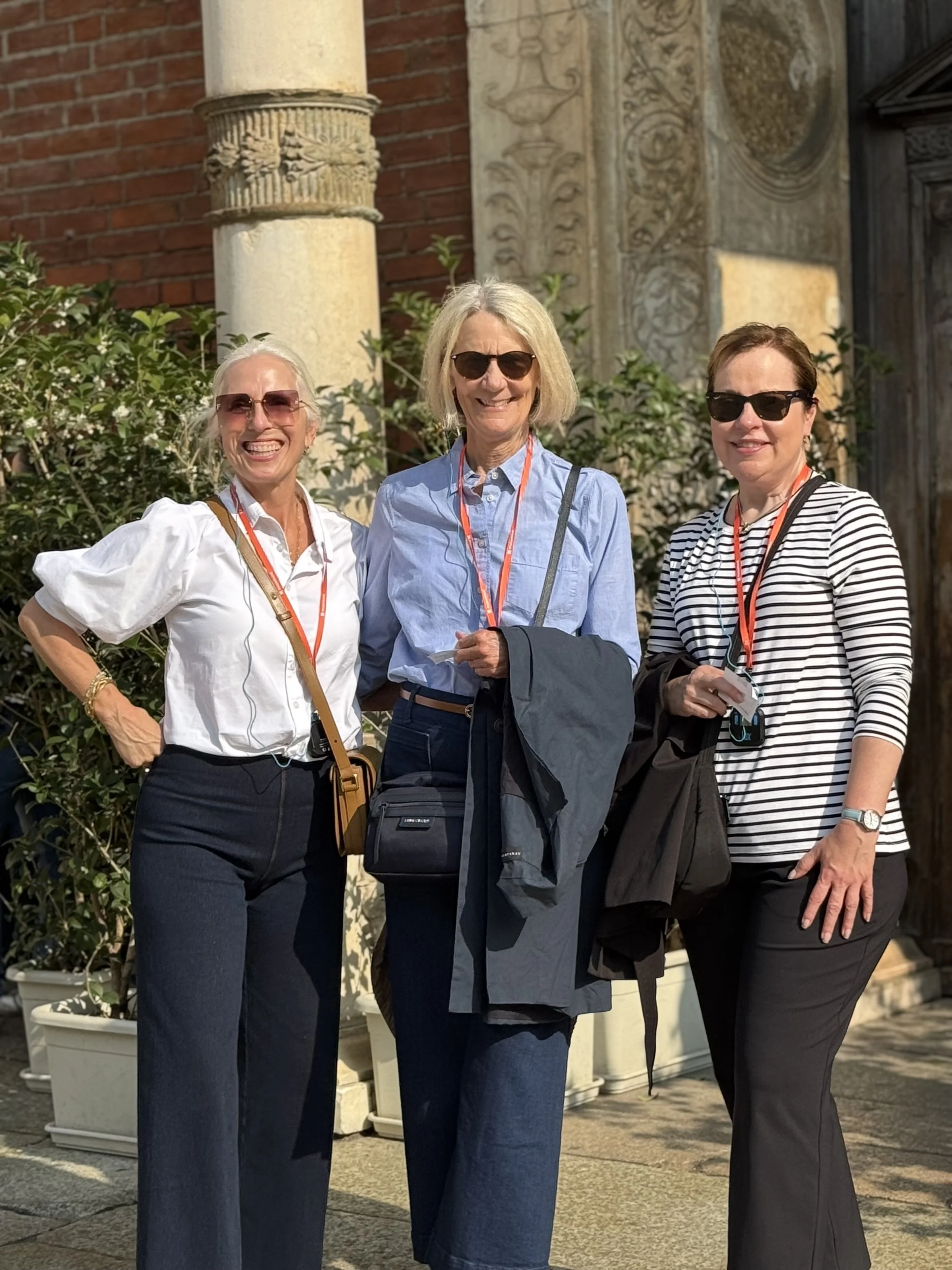 Three women standing outdoors in front of a historic building, smiling and posing for a photo on a sunny day, wearing sunglasses and casual professional clothing.