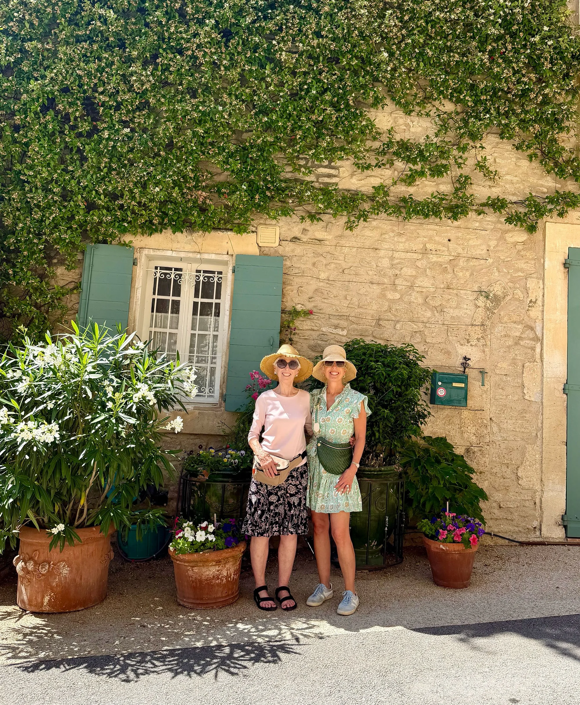 Two women wearing large sun hats and sunglasses standing in front of a stone wall covered with climbing plants and white flowers, with potted plants and flowers at their feet.