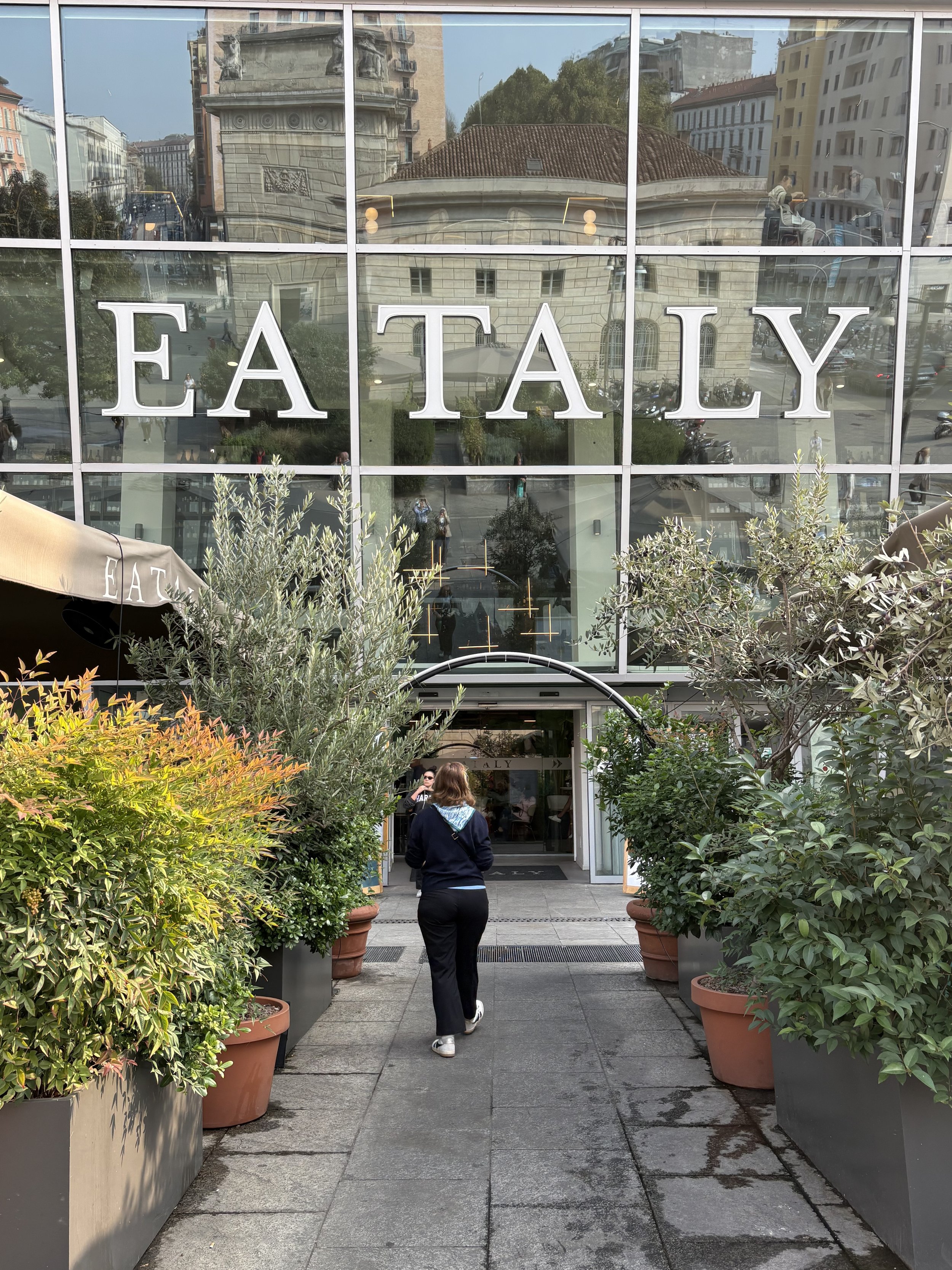 People entering a building labeled EATALY, with large potted plants on either side of the walkway.
