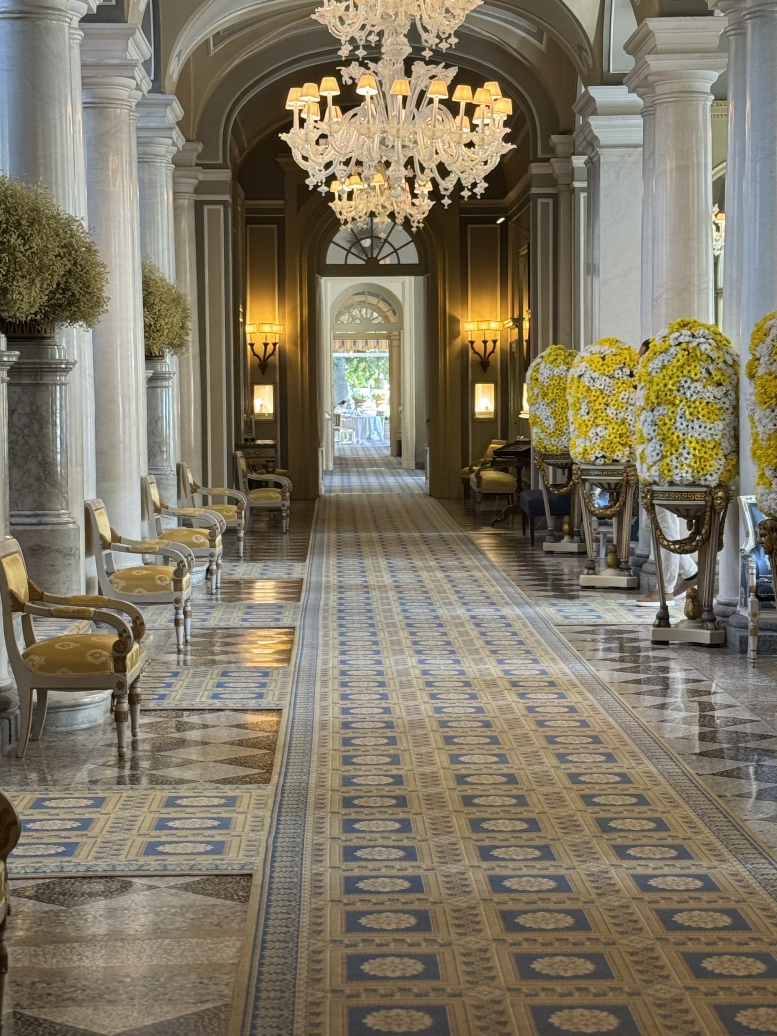 Elegant hallway with marble columns, ornate chandeliers, and yellow and white floral arrangements.