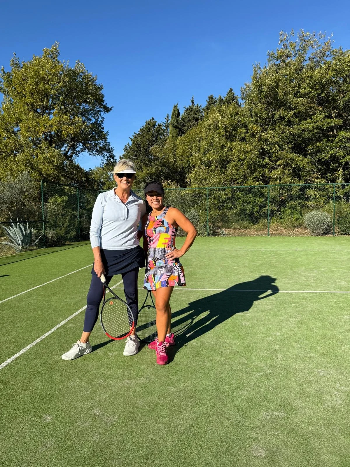 Two women standing on a tennis court, holding tennis rackets, smiling, with trees and a blue sky in the background.