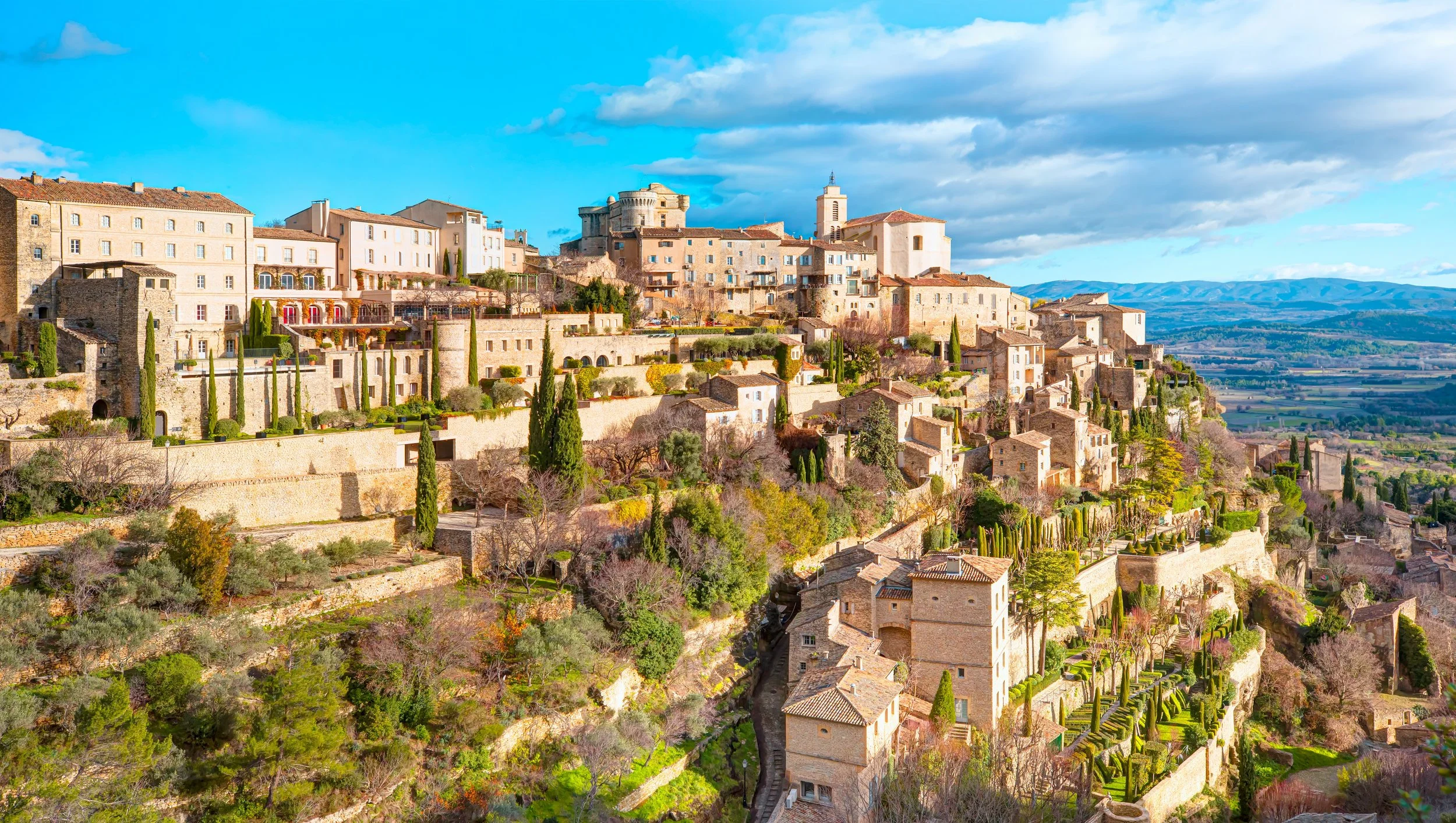 A hillside town with stone buildings, trees, and terraced gardens under a partly cloudy sky.