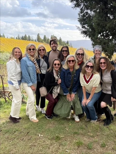 Group of ten women and one man posing outdoors in front of yellow flowering plants, with trees and cloudy sky in the background.