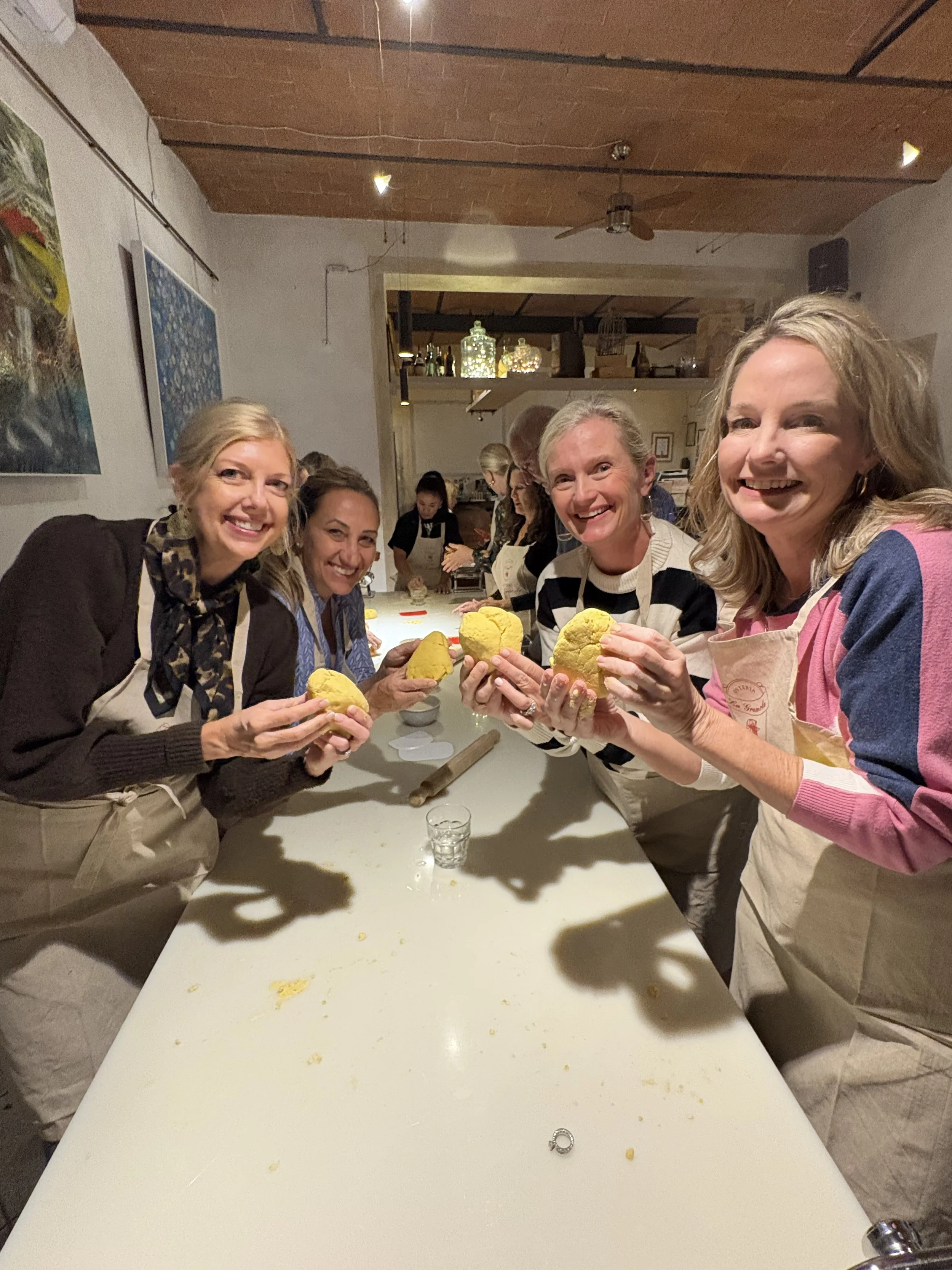 Four women smiling and holding yellow baking cookies in a kitchen or bakery setting.