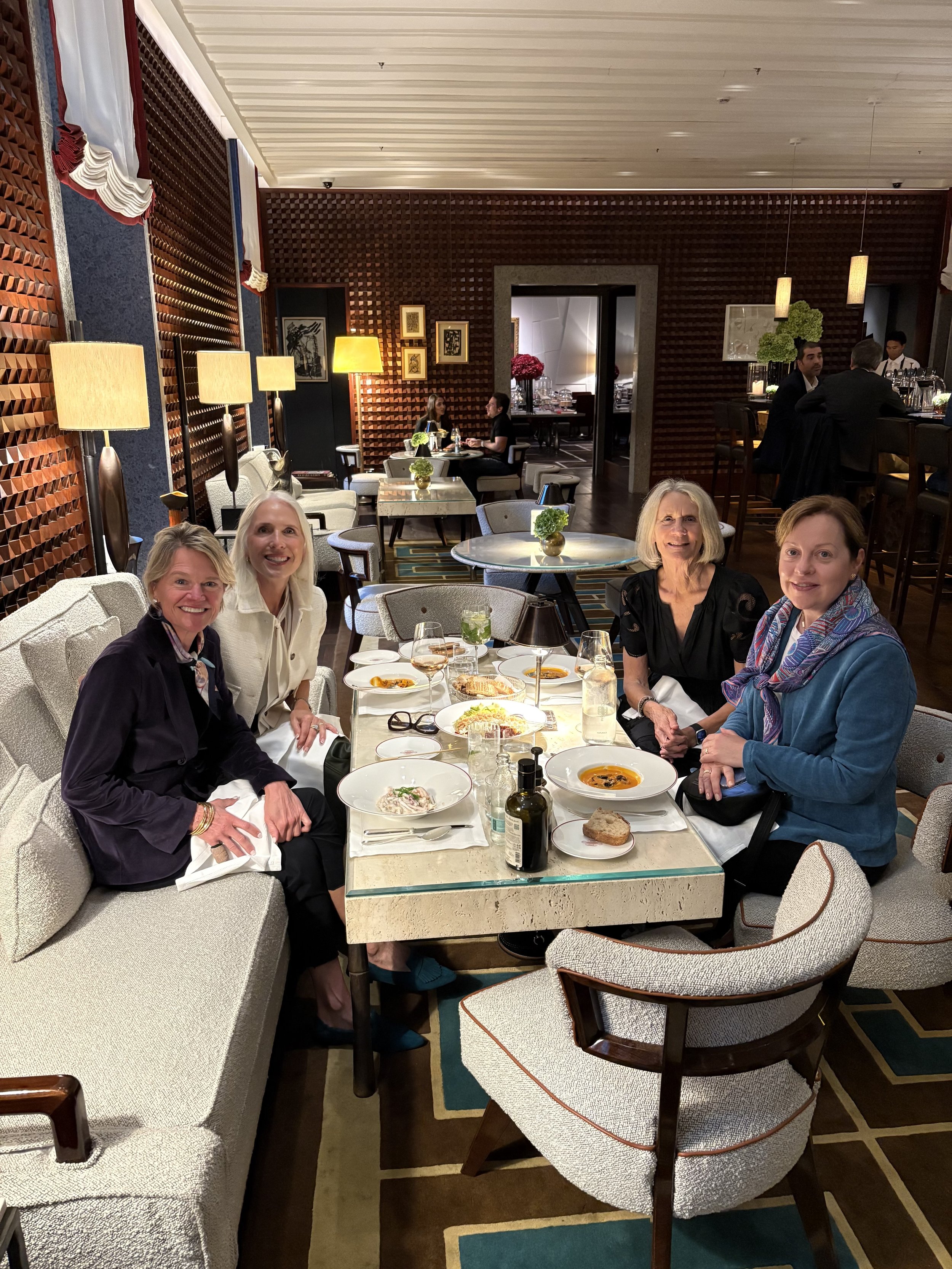 Four women sitting at a dining table in a restaurant, with plates of soup, bread, salads, and drinks, smiling at the camera.