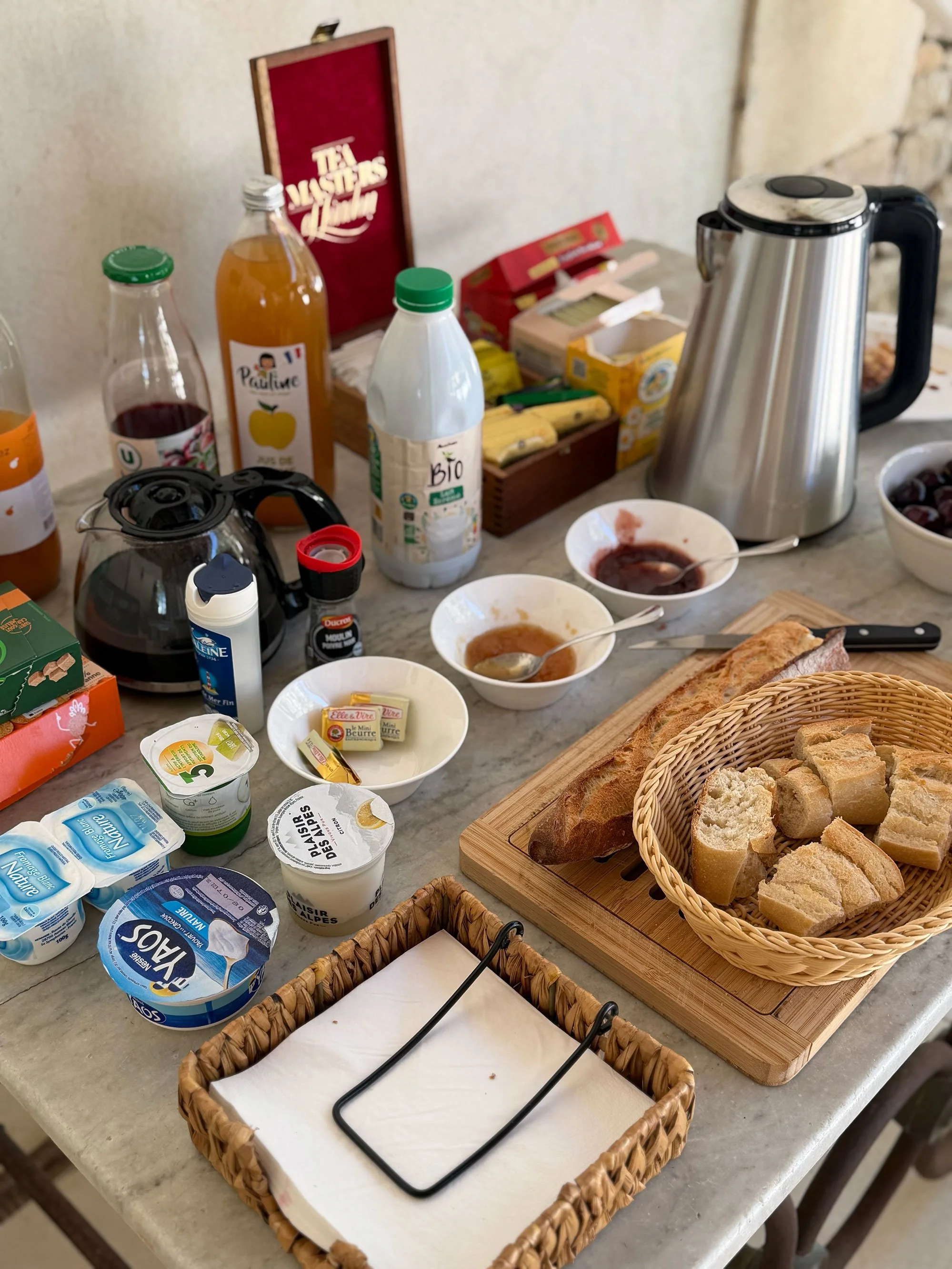 A breakfast table with various bread, yogurt, and fruit, along with a thermal carafe, juice bottles, and small bowls with jam or compote and a spoon.
