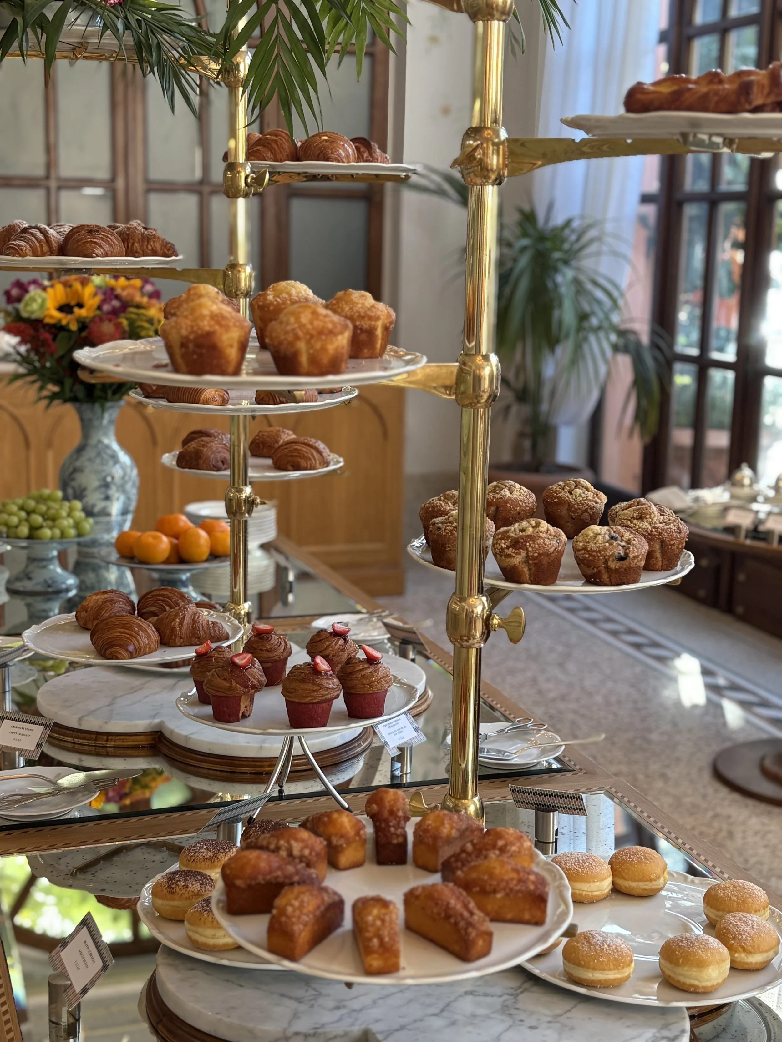 A multi-tiered display stand filled with various pastries, muffins, and cupcakes in a room with large windows, a wooden cabinet, and a bouquet of colorful flowers.