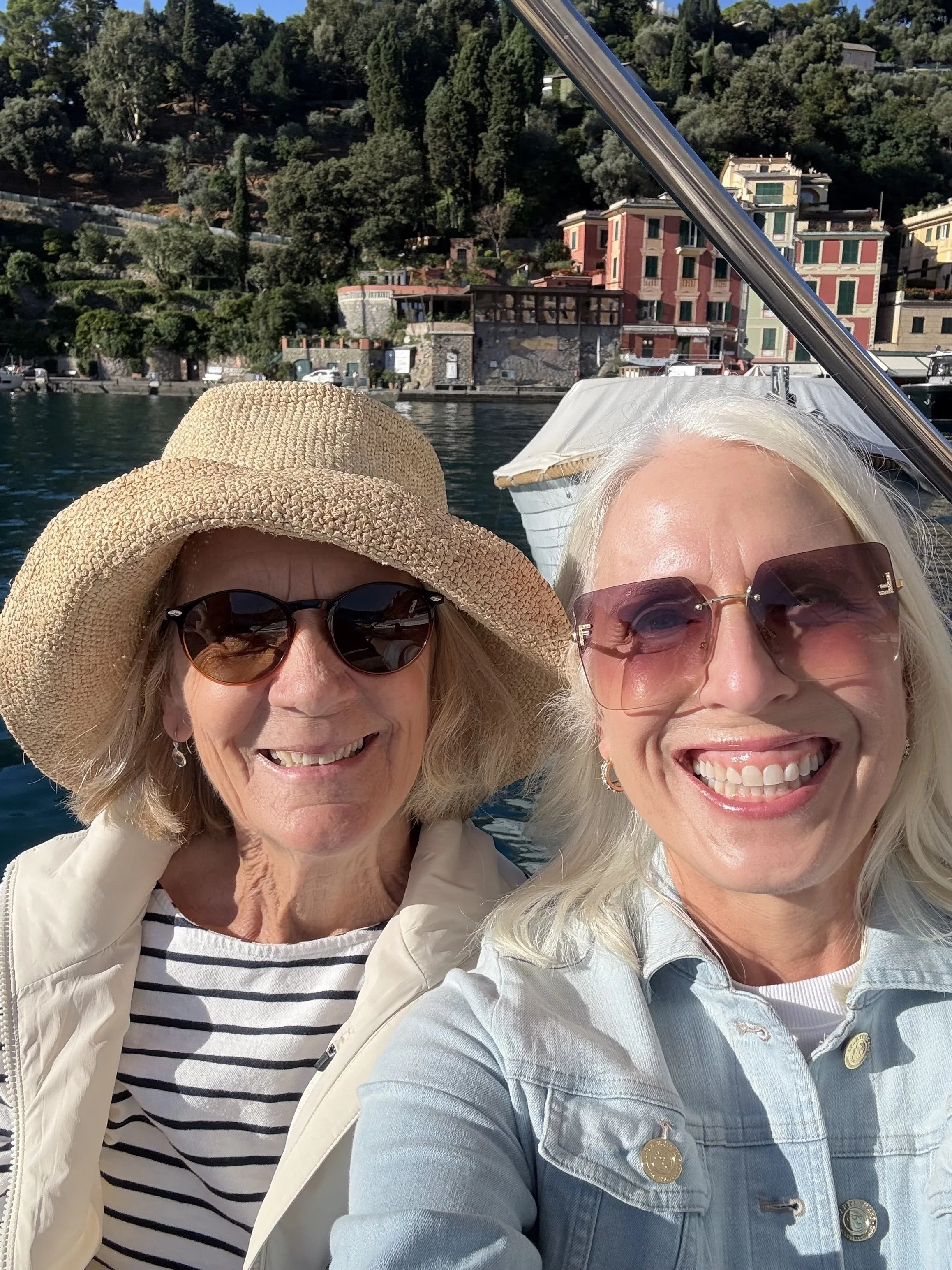 Two women taking a selfie on a boat with water, hillside, and colorful buildings in the background.