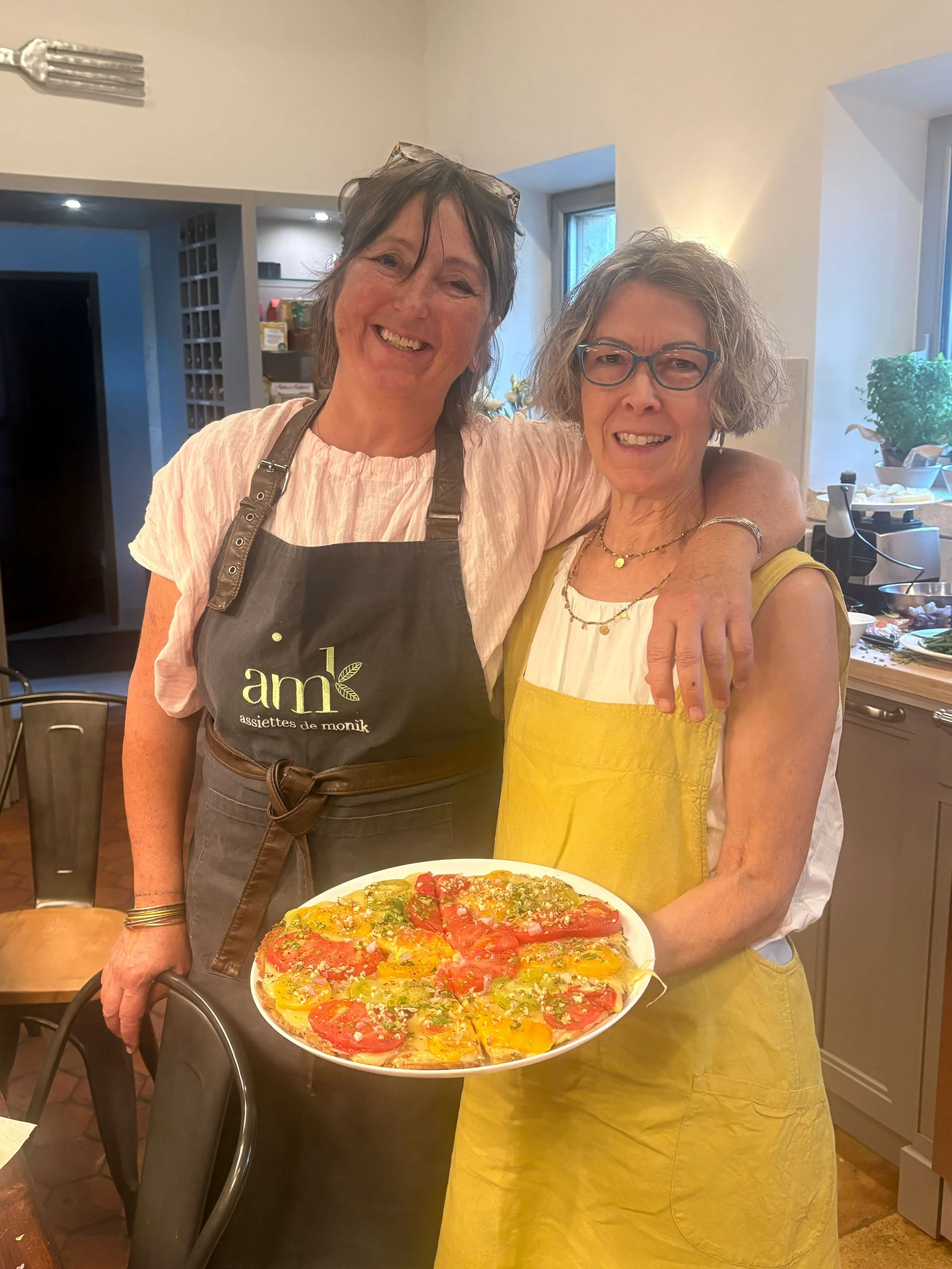 Two smiling women standing in a kitchen, one wearing a black apron holding a colorful pizza, the other wearing glasses and a yellow apron.