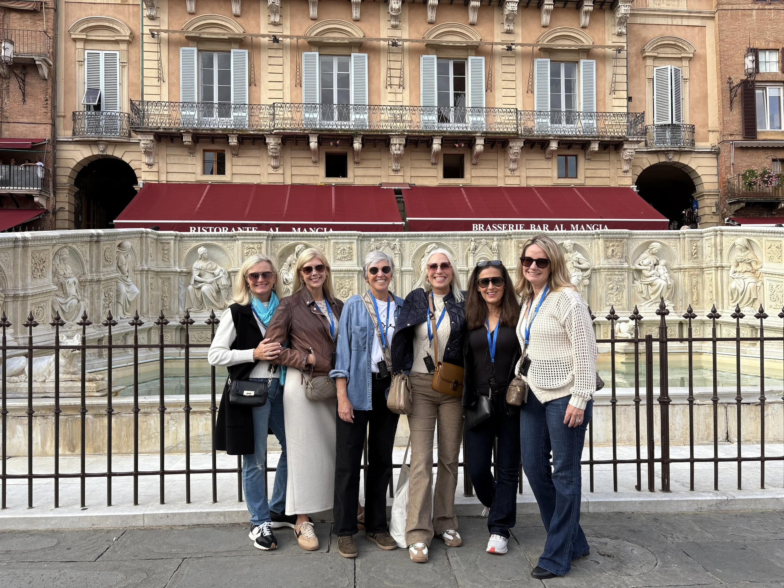 A group of six women standing in front of a fountain and ornate building in a city, wearing casual clothing and sunglasses.
