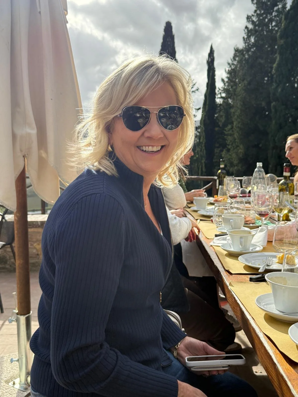 Smiling blonde woman wearing sunglasses and a navy blue jacket sitting at an outdoor dining table with empty plates, glasses, and a woman with dark hair in the background, trees and cloudy sky visible.