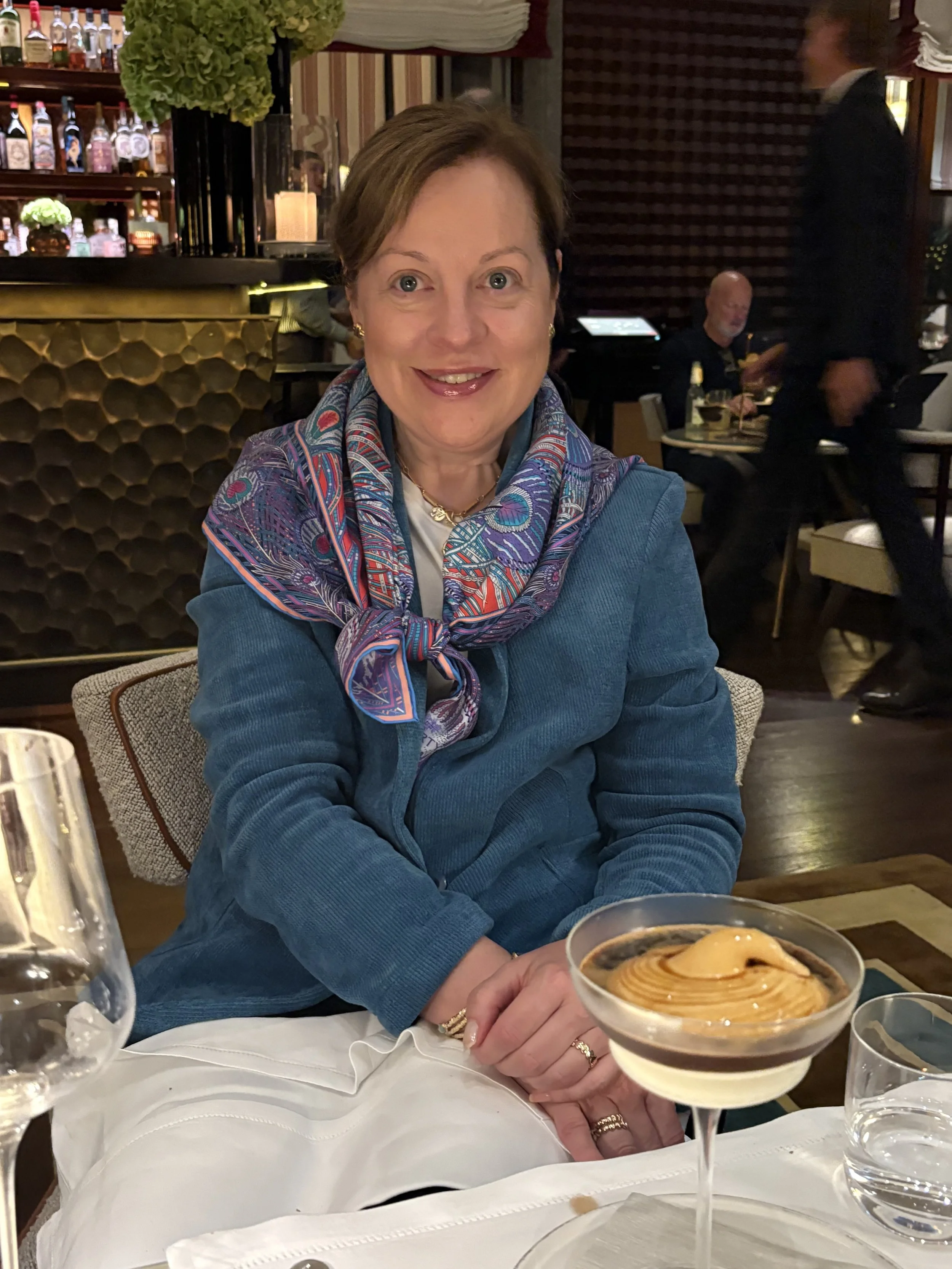 A woman sitting at a dining table in a restaurant, smiling, with a dessert and drinks on the table, and waitstaff serving in the background.