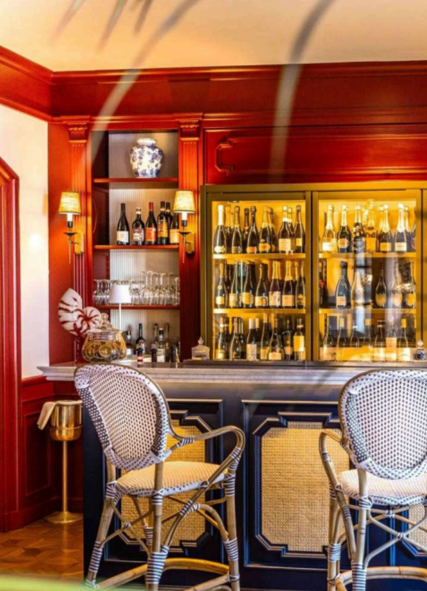 A bar with a dark wood counter, two rattan bar stools with woven seats, a glass display case with champagne bottles, wine bottles and glasses behind the bar, and decorative lighting and shelves with a vase and additional bottles.