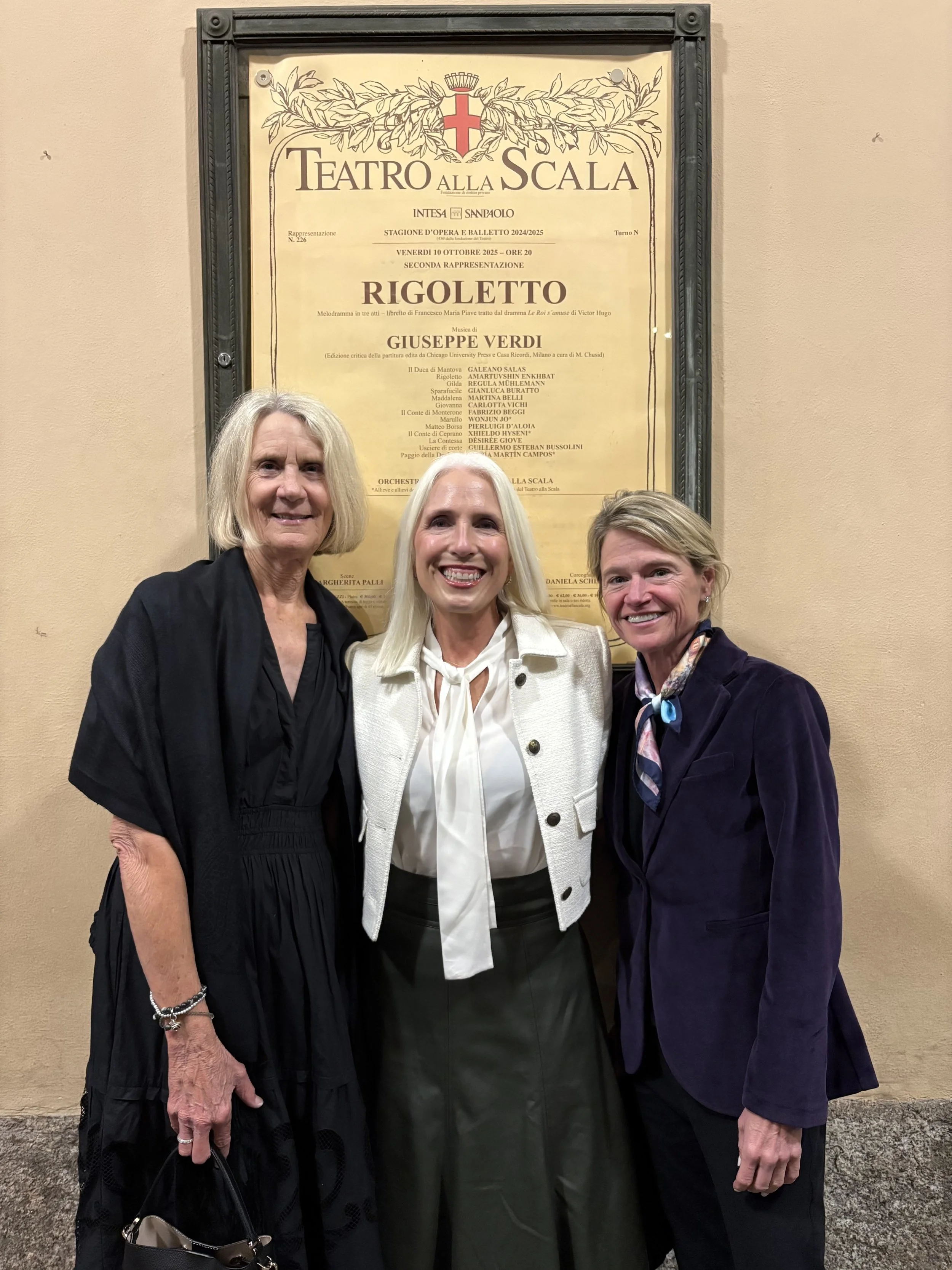 Three women smiling in front of a yellow theater poster for Teatro Alla Scala.
