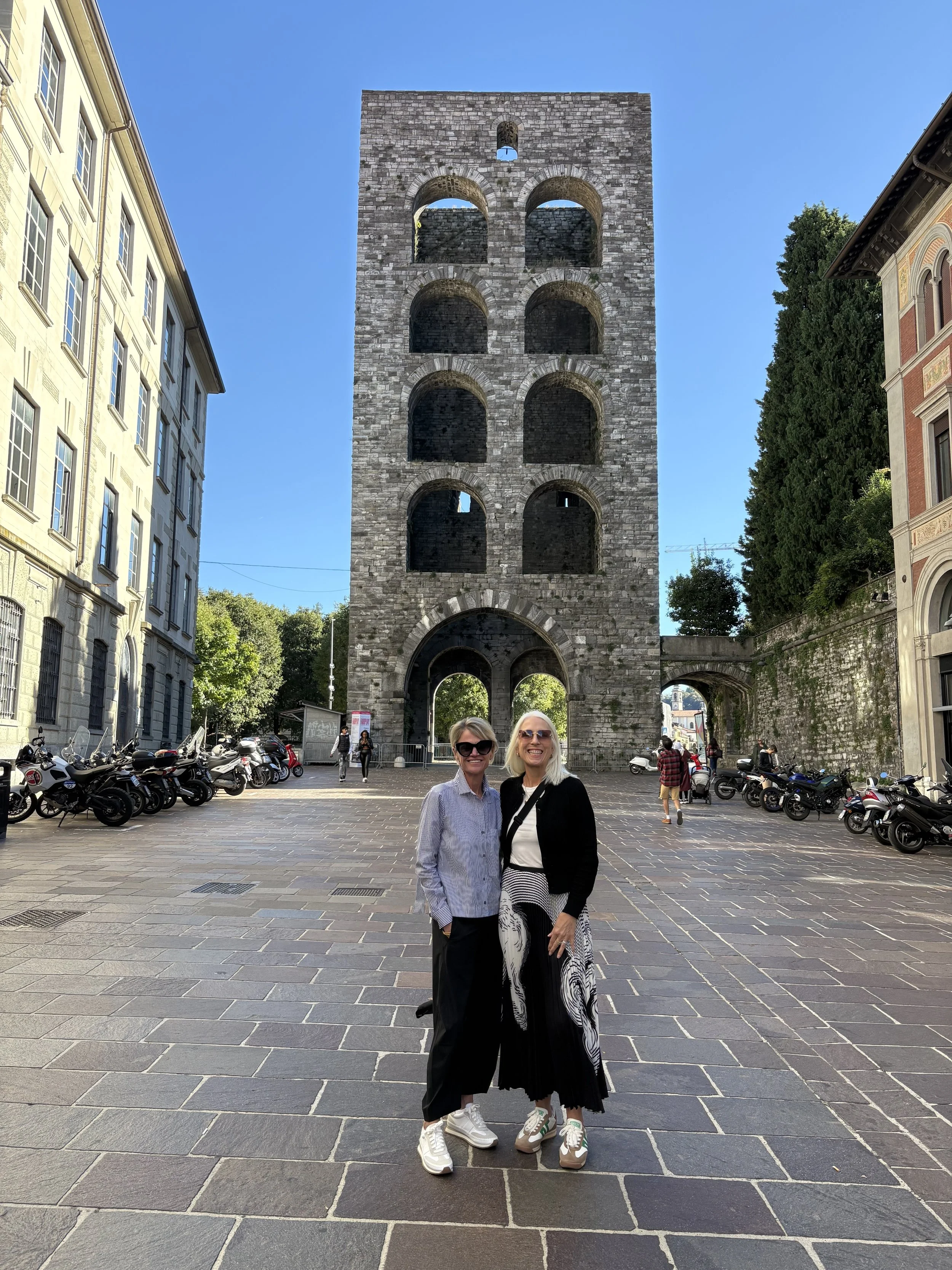 Two women standing in front of the medieval Torre dei Lamberti clock tower in Verona, Italy, smiling and posing for a photo.