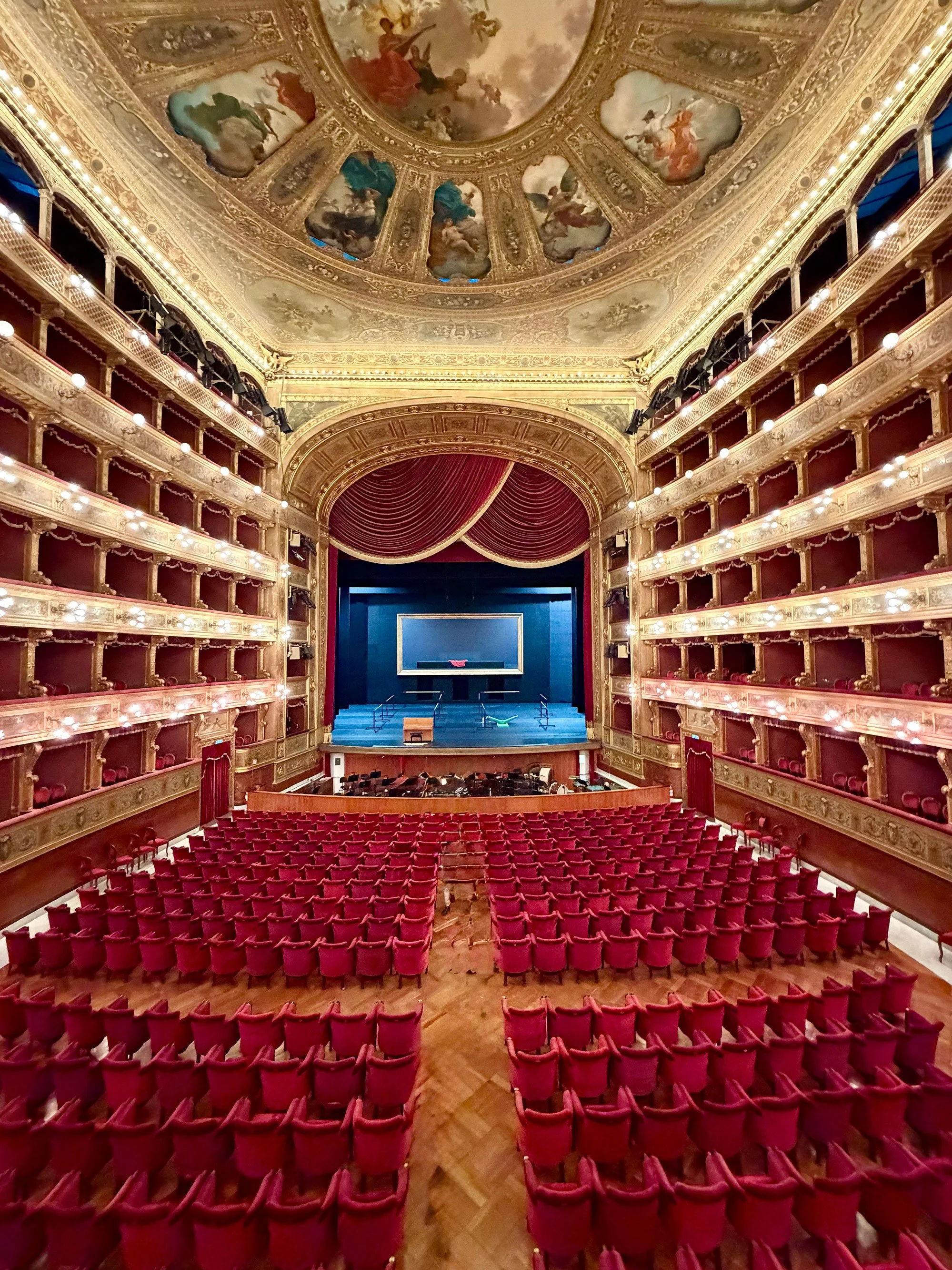Empty ornate theater with red velvet seats, decorated gold accents, and a stage with red curtains.