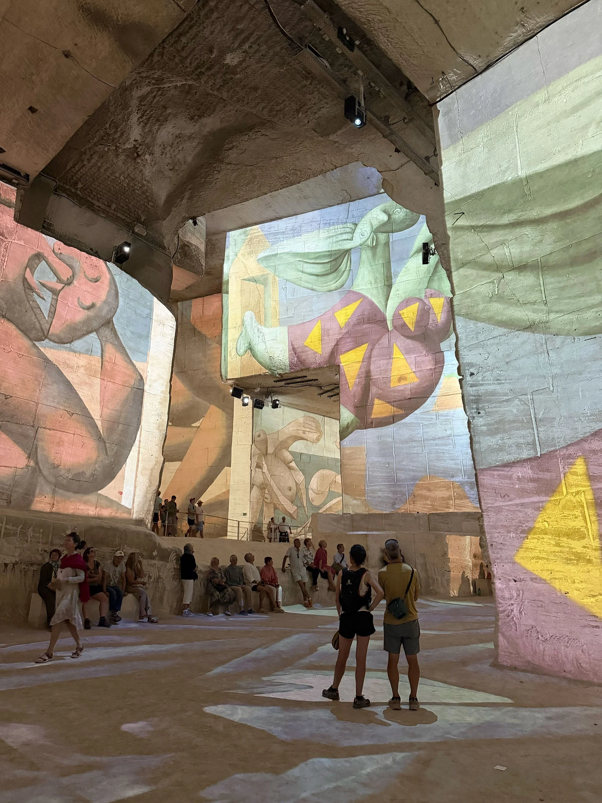 An underground venue with stone walls and ceiling, featuring large wall projections or murals of abstract and mythical figures. People are seated and standing, observing the art exhibition.